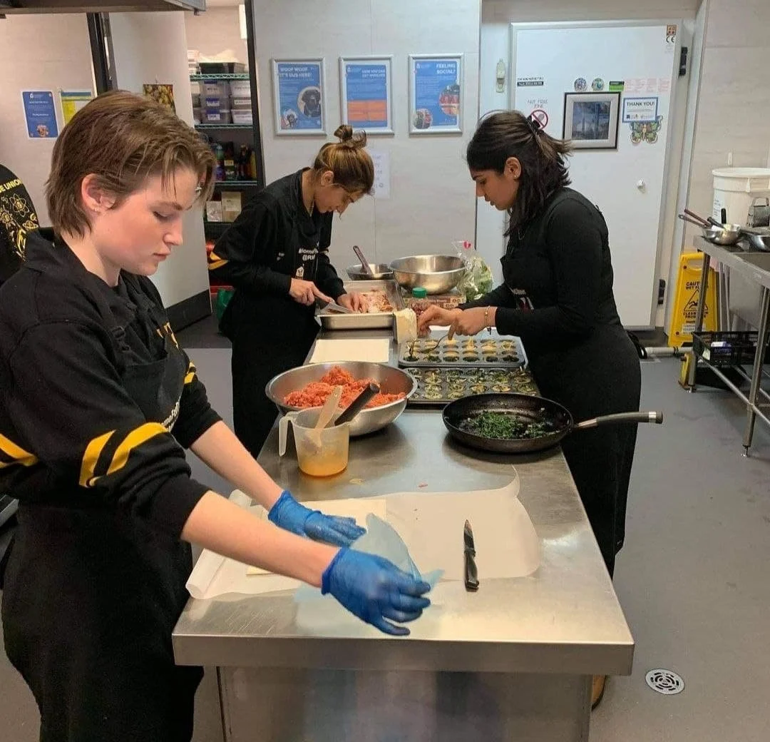 Three women in black uniforms preparing food in a kitchen. The woman at the front is wearing blue gloves and handling dough at a stainless steel counter. The other two women are working with ingredients on their respective prep stations.