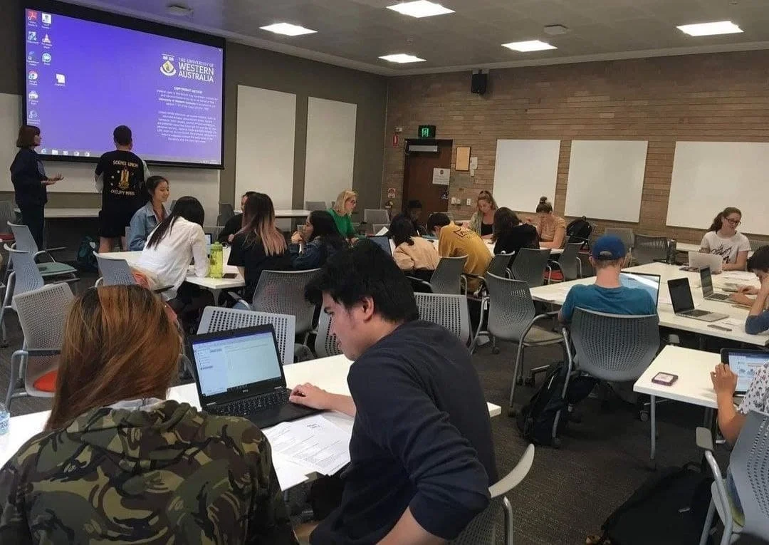 A classroom or lecture hall at The University of Western Australia filled with students studying and working on laptops. There is a large screen displaying the university's logo and information at the front of the room.