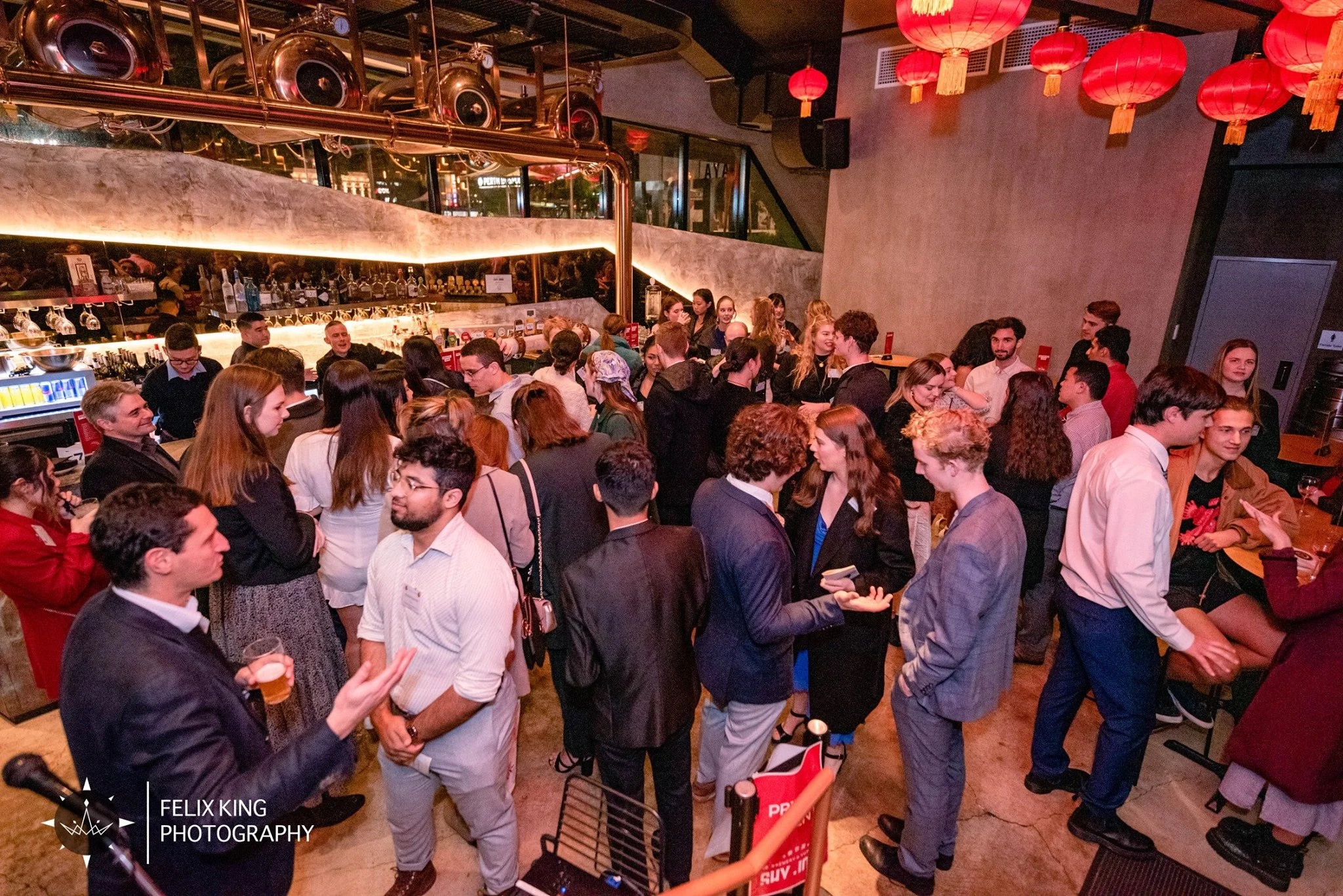 Crowd of people at a bar or nightclub with red lanterns hanging from the ceiling, a bar counter in the background, and various drinks and bar equipment on display.