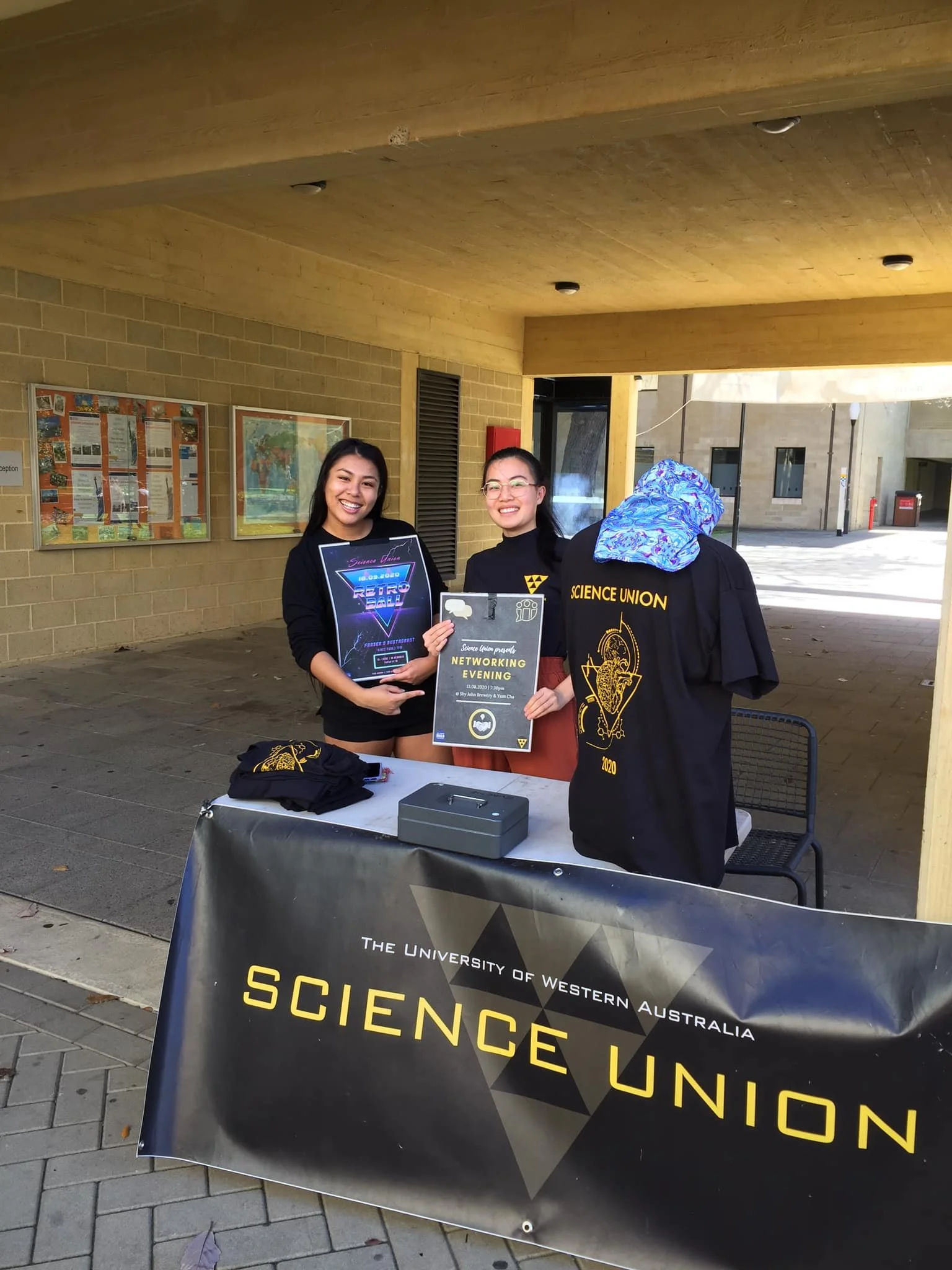 Three young women standing behind a table at a Science Union event, smiling and holding a poster, with a banner displaying 'The University of Western Australia Science Union' in front of them.