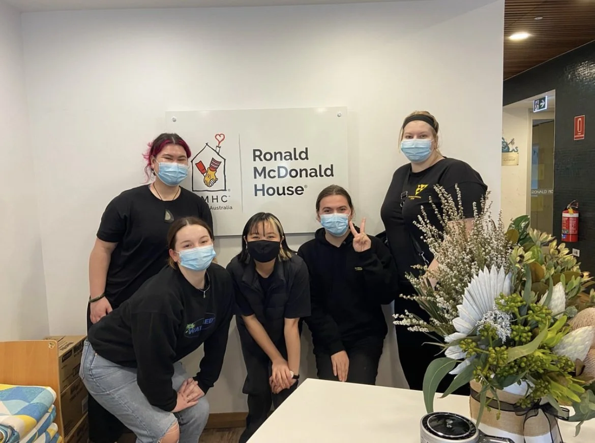 Group of five women wearing masks standing and sitting in front of a Ronald McDonald House sign, with a bouquet of flowers on a table in the foreground.