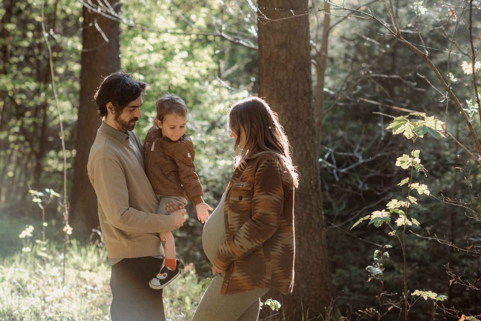 Pregnant mother standing with her partner and child in soft forest light during a relaxed family maternity session.