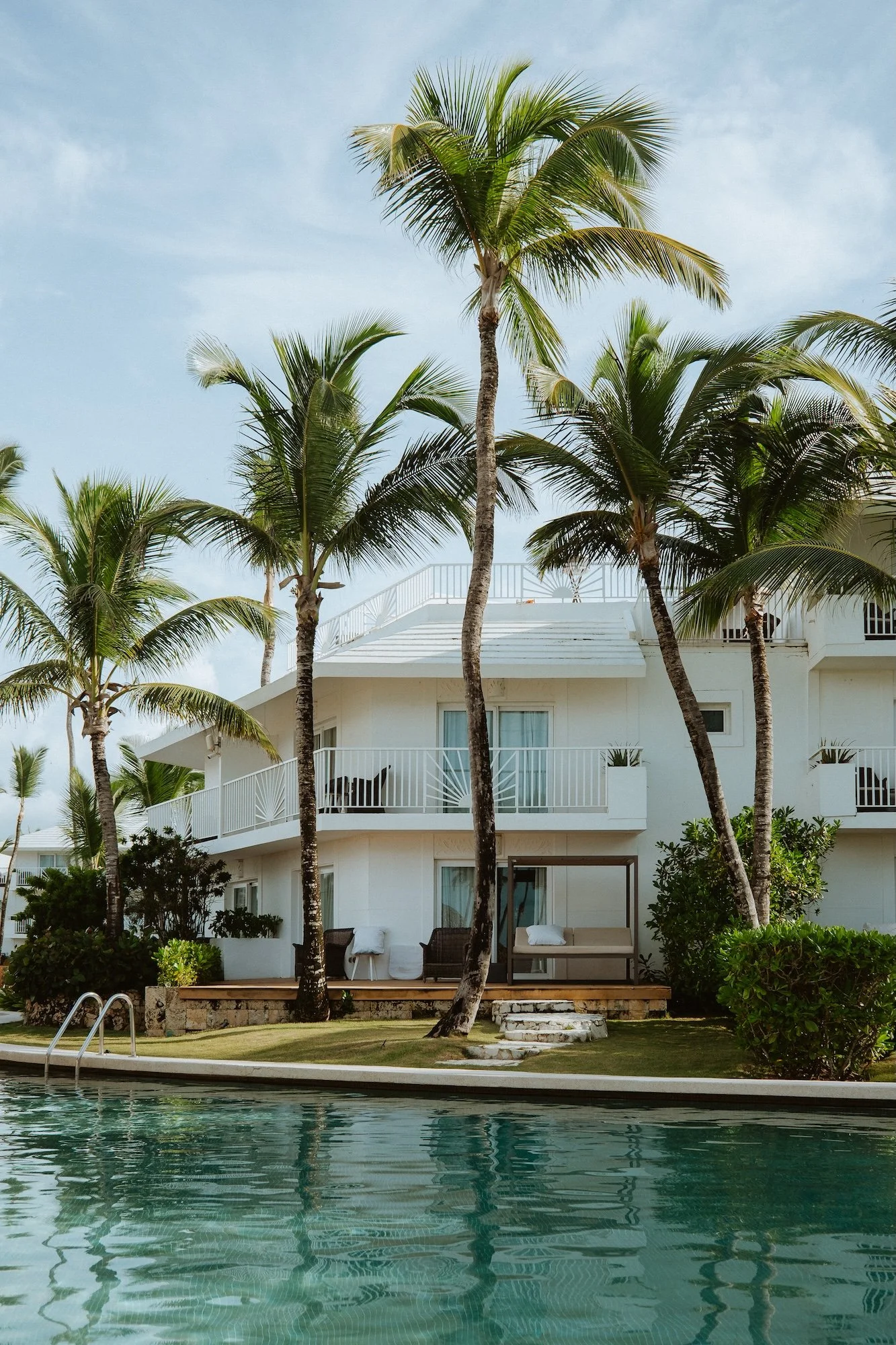 A tropical resort with a swimming pool, white building with balconies, and tall palm trees under a partly cloudy sky.
