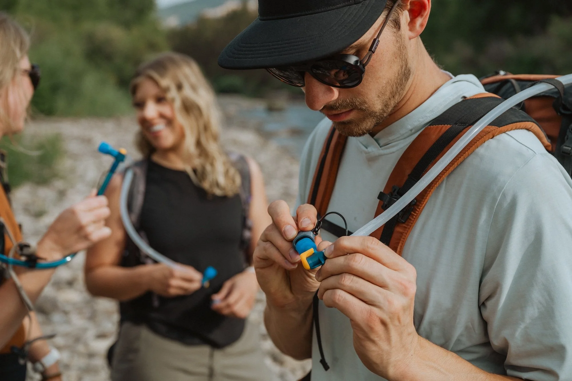 Three hikers outdoors, one man adjusting gear with a backpack, the two women smiling and holding equipment, in a natural setting with trees and rocks.