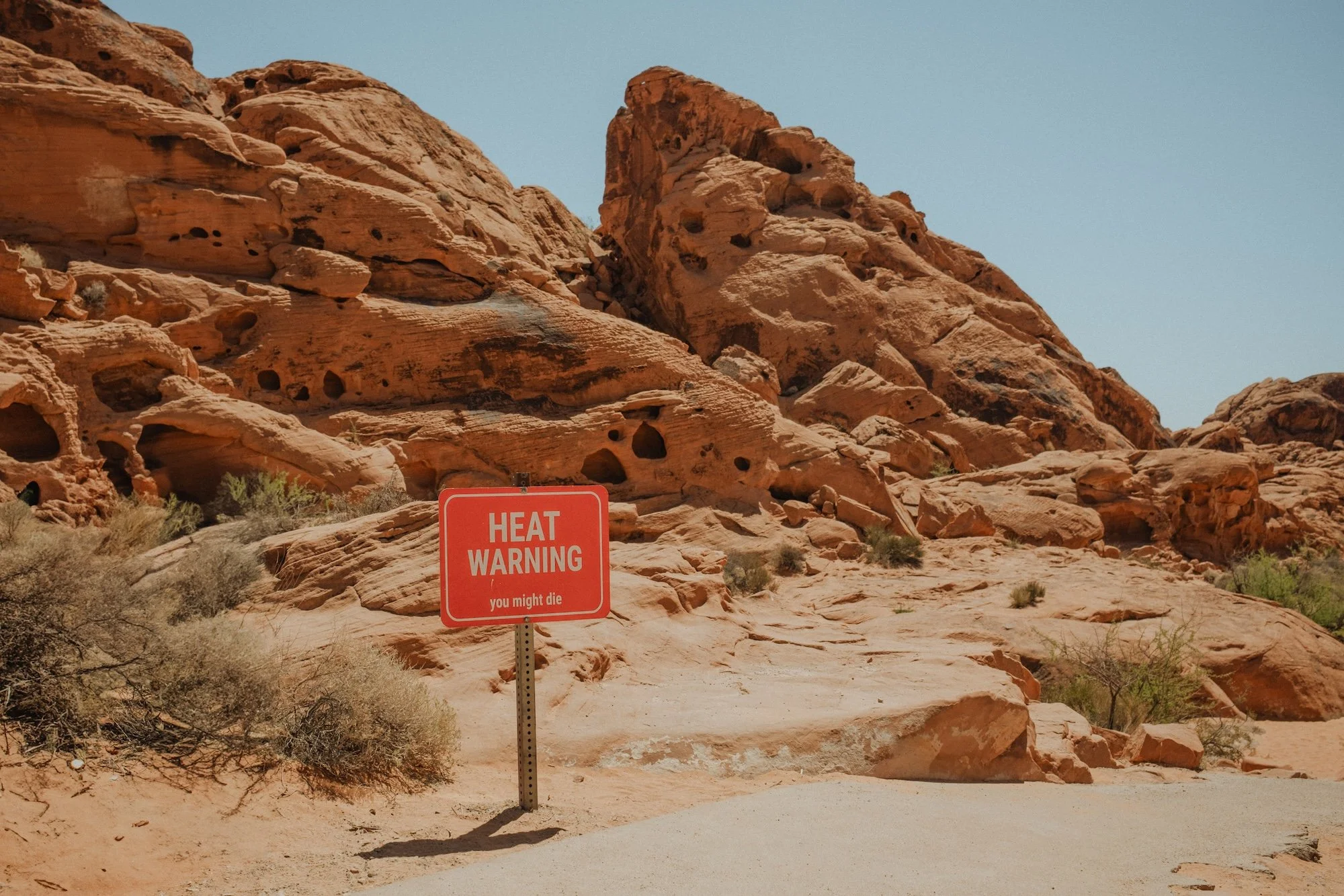 Red heat warning sign in desert with rocky formations and sparse vegetation under clear blue sky.