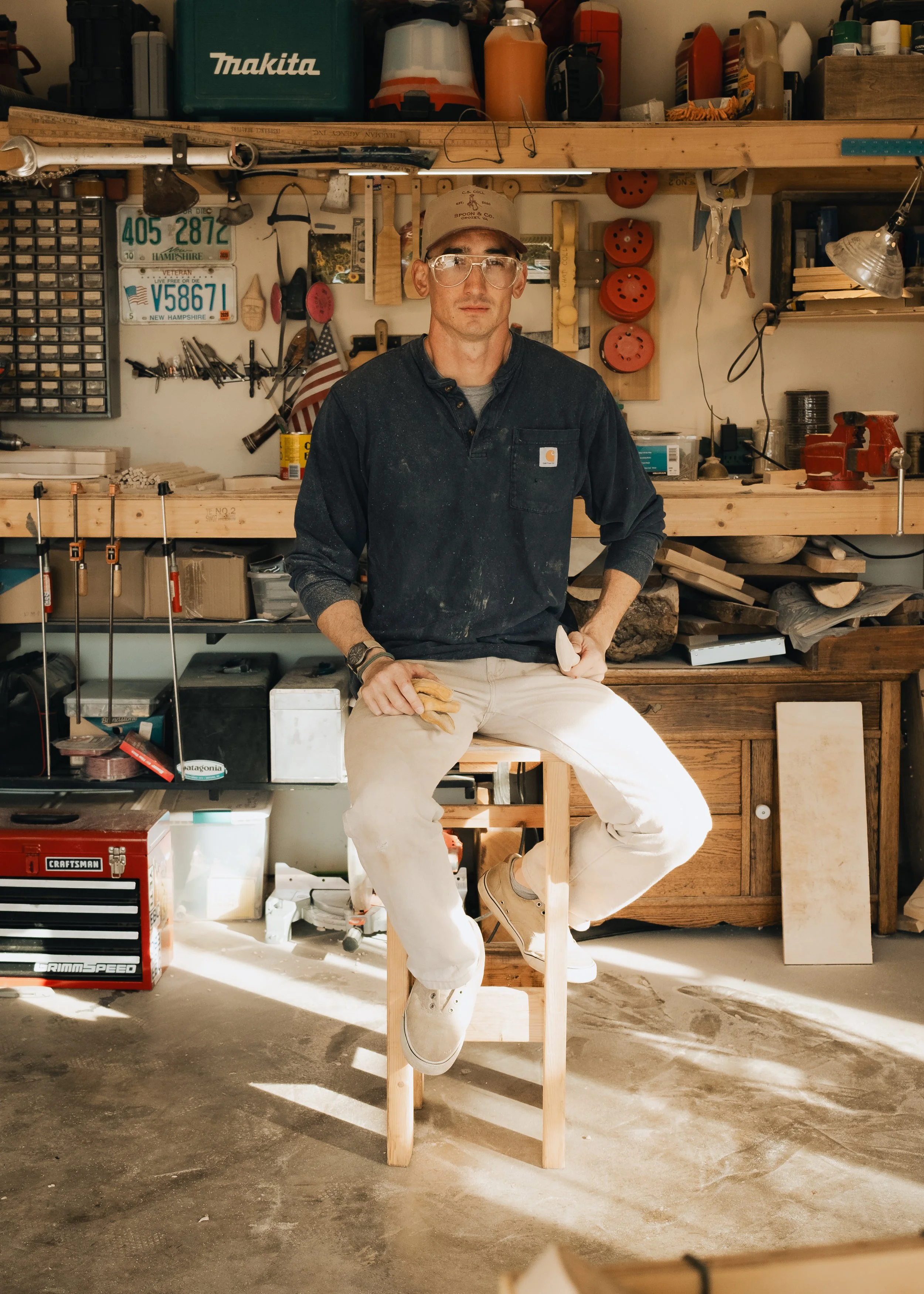 A man sitting on a wooden stool in a woodworking workshop, surrounded by tools and equipment, wearing glasses, a cap, and a dark long-sleeve shirt.