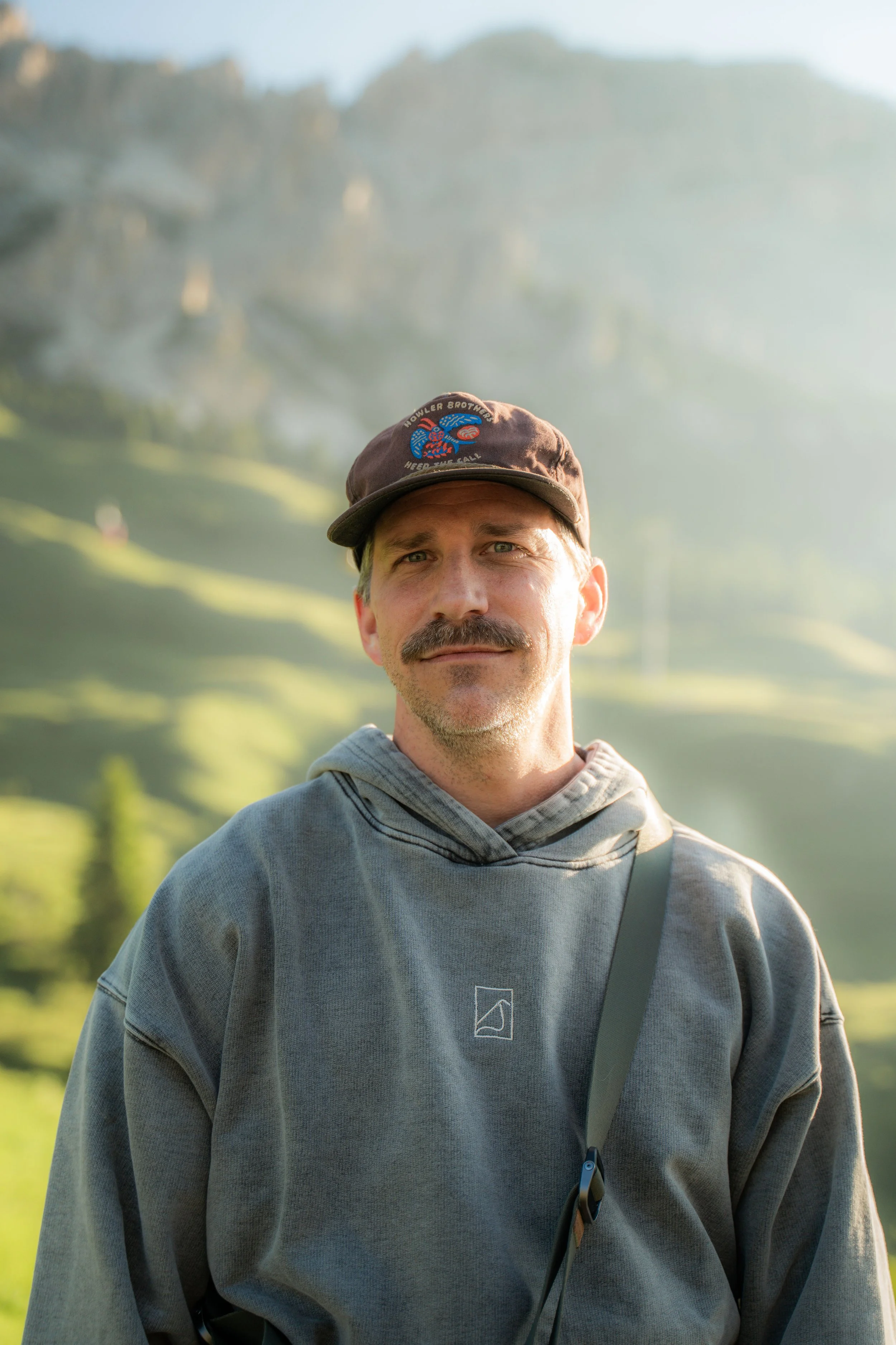 A man with a mustache wearing a grey hoodie and a brown baseball cap with patches, standing outdoors with a mountainous landscape and trees in the background.