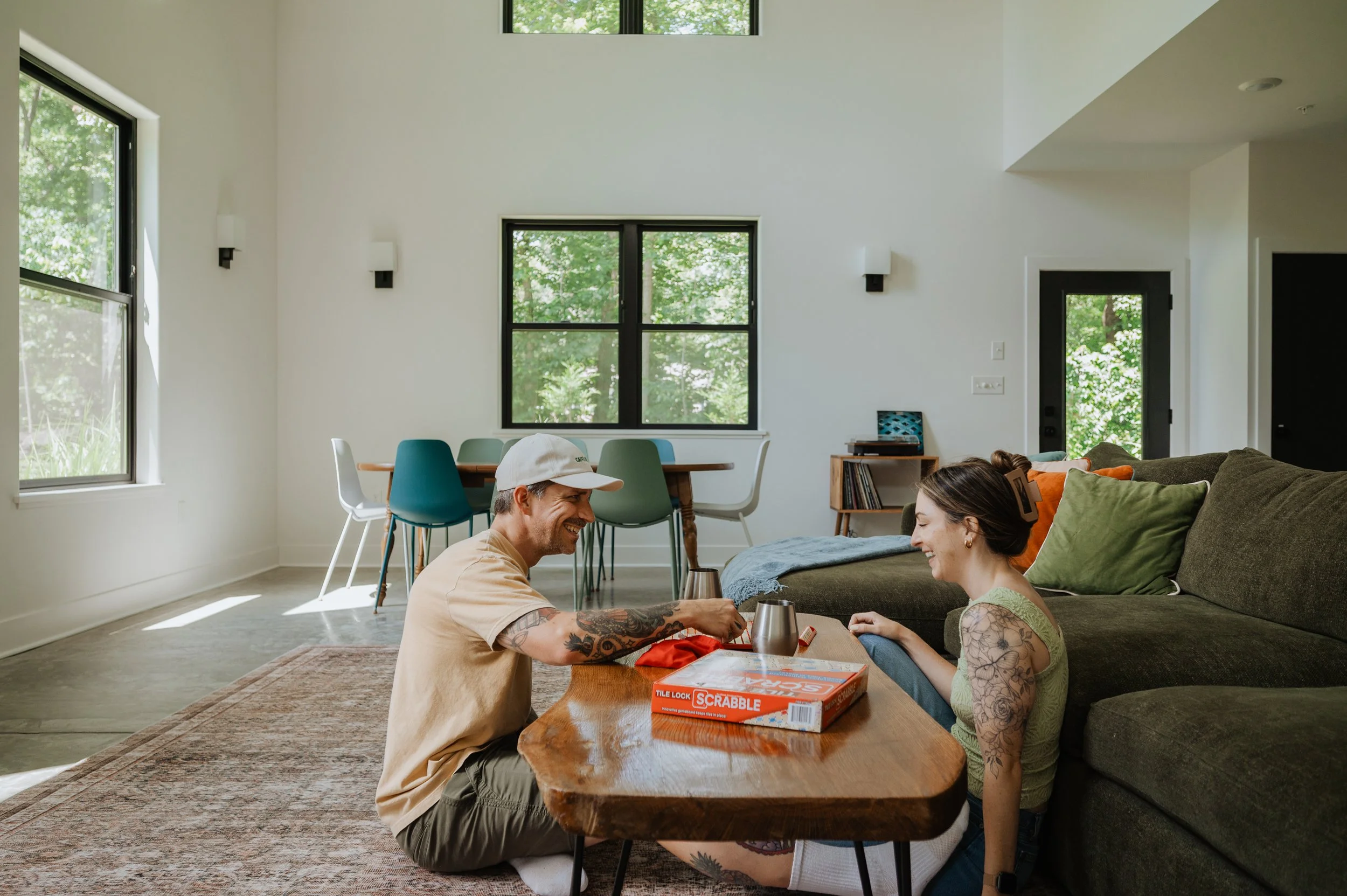 A man and woman playing Scrabble at a coffee table in a modern living room with large windows and a dining area in the background.