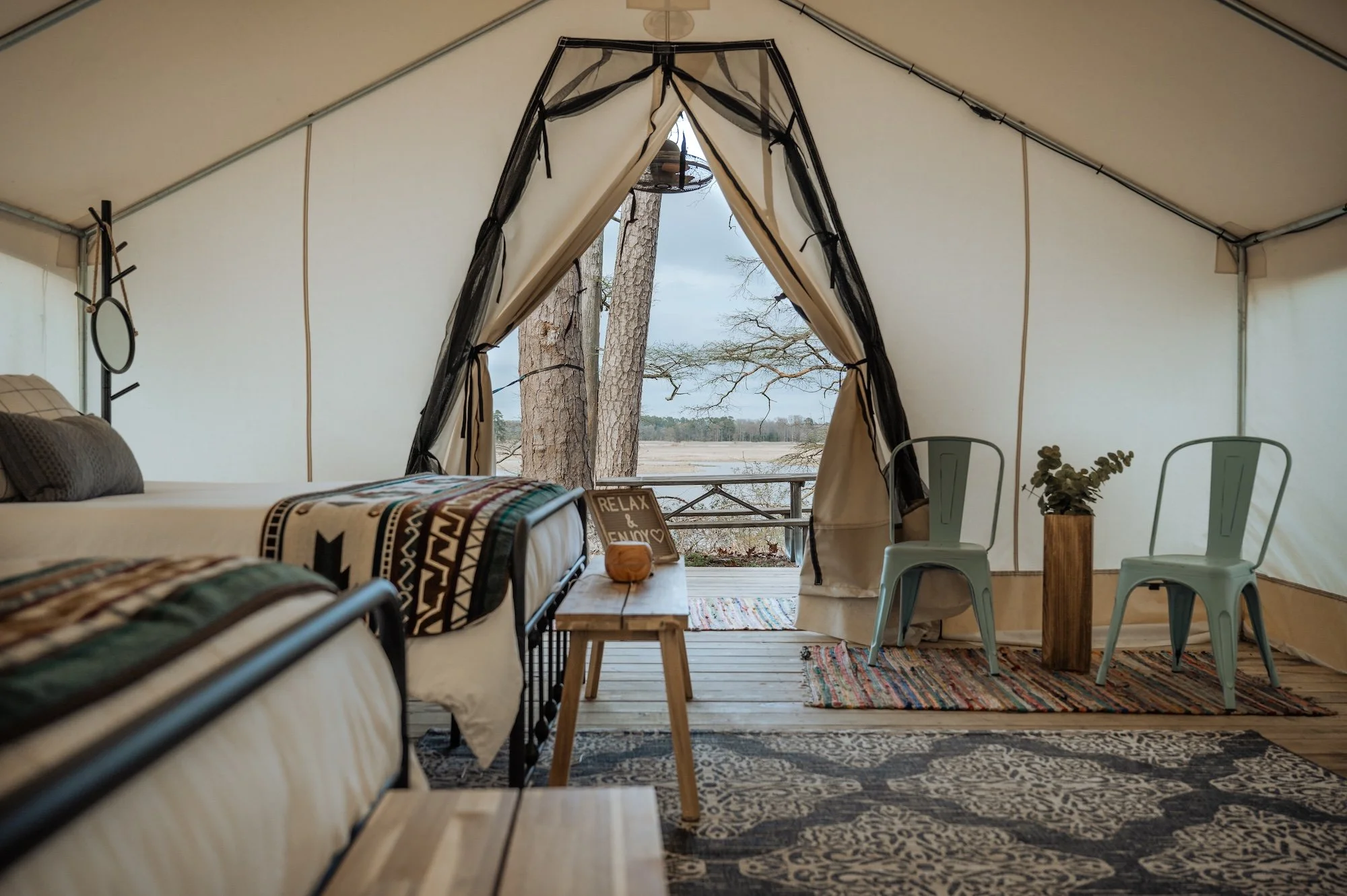 Interior view of a cozy glamping tent with two beds, a small wooden table, two metal chairs, and a view of trees outside through the open tent entrance.