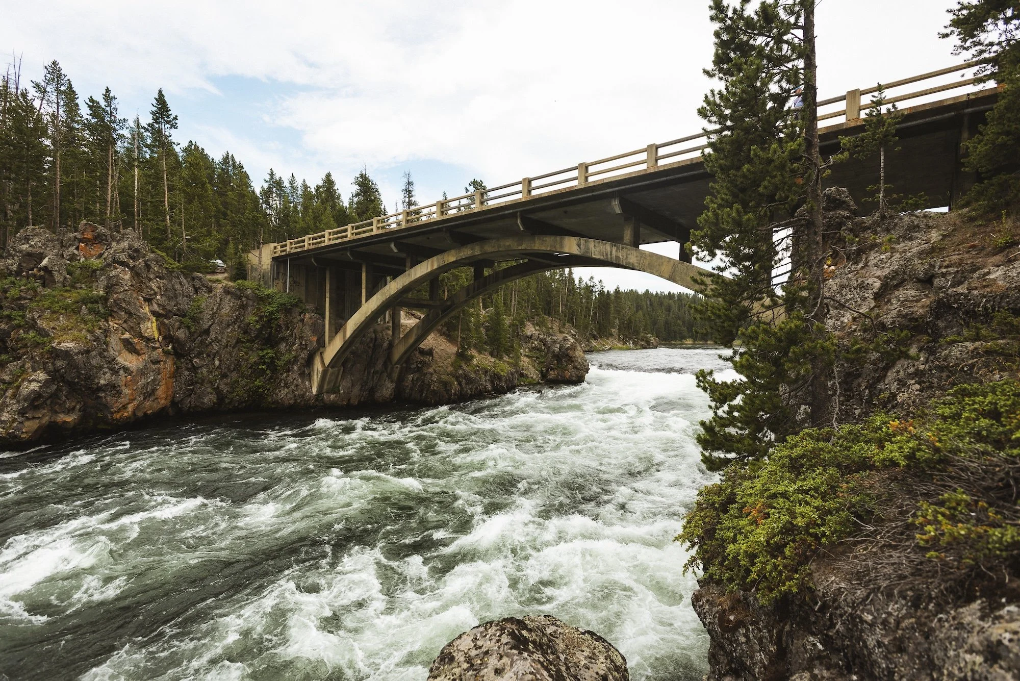 View of a bridge over a fast-moving river in a forested canyon.