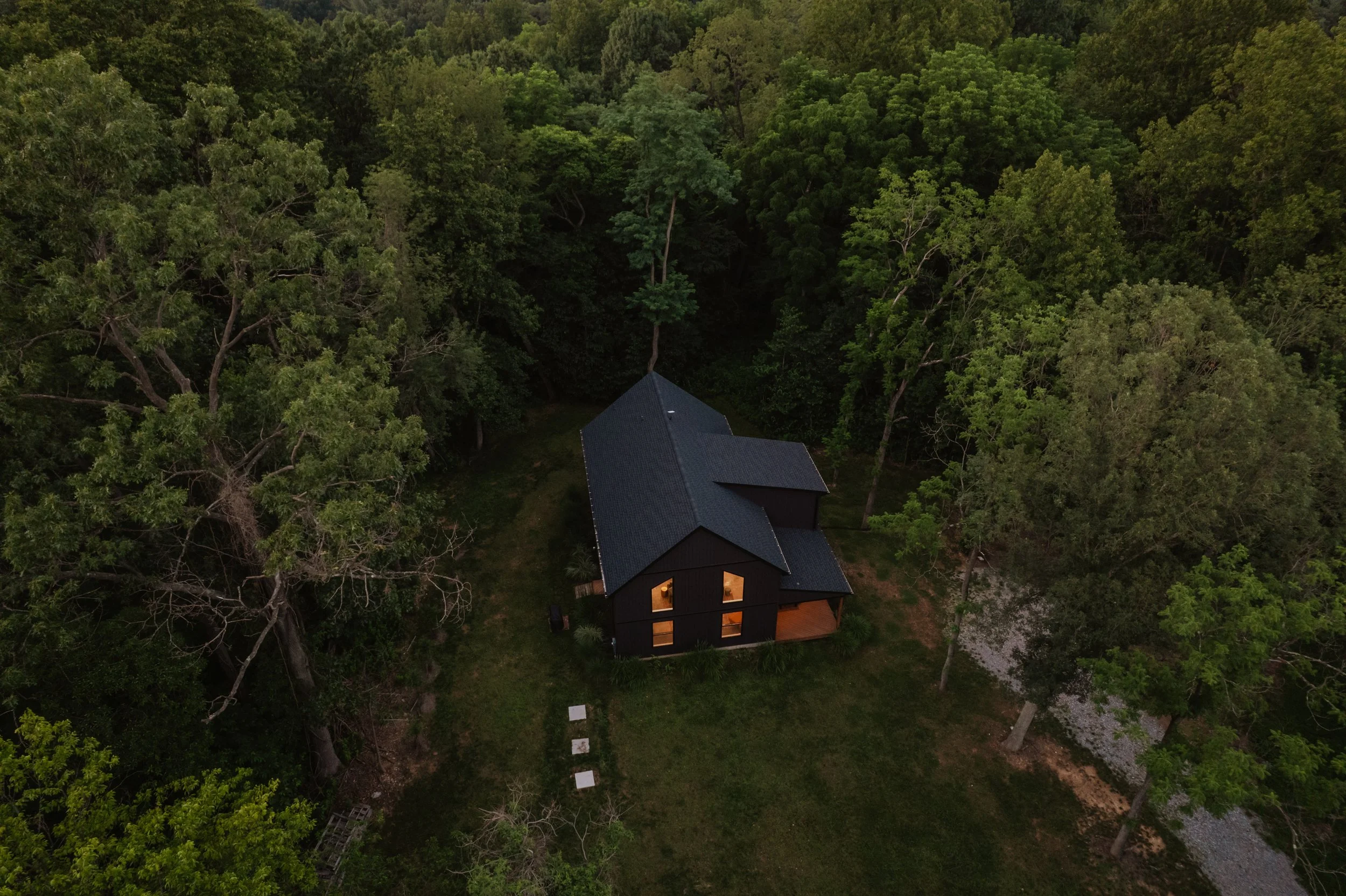 A black house with lights on inside, surrounded by green trees, viewed from above at dusk.