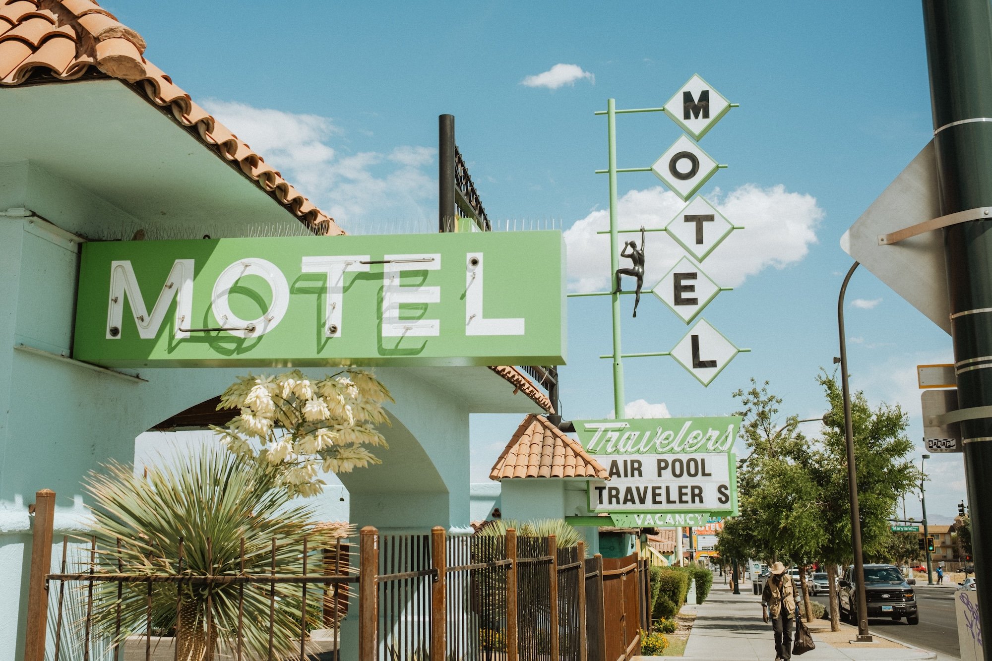 Street view of a motel with a large green sign that says 'MOTEL', and a vertical sign spelling 'MOTEL' with a small sculpture of a woman hanging from the letter 'T'. There are trees, a fence, and pedestrians on the sidewalk.