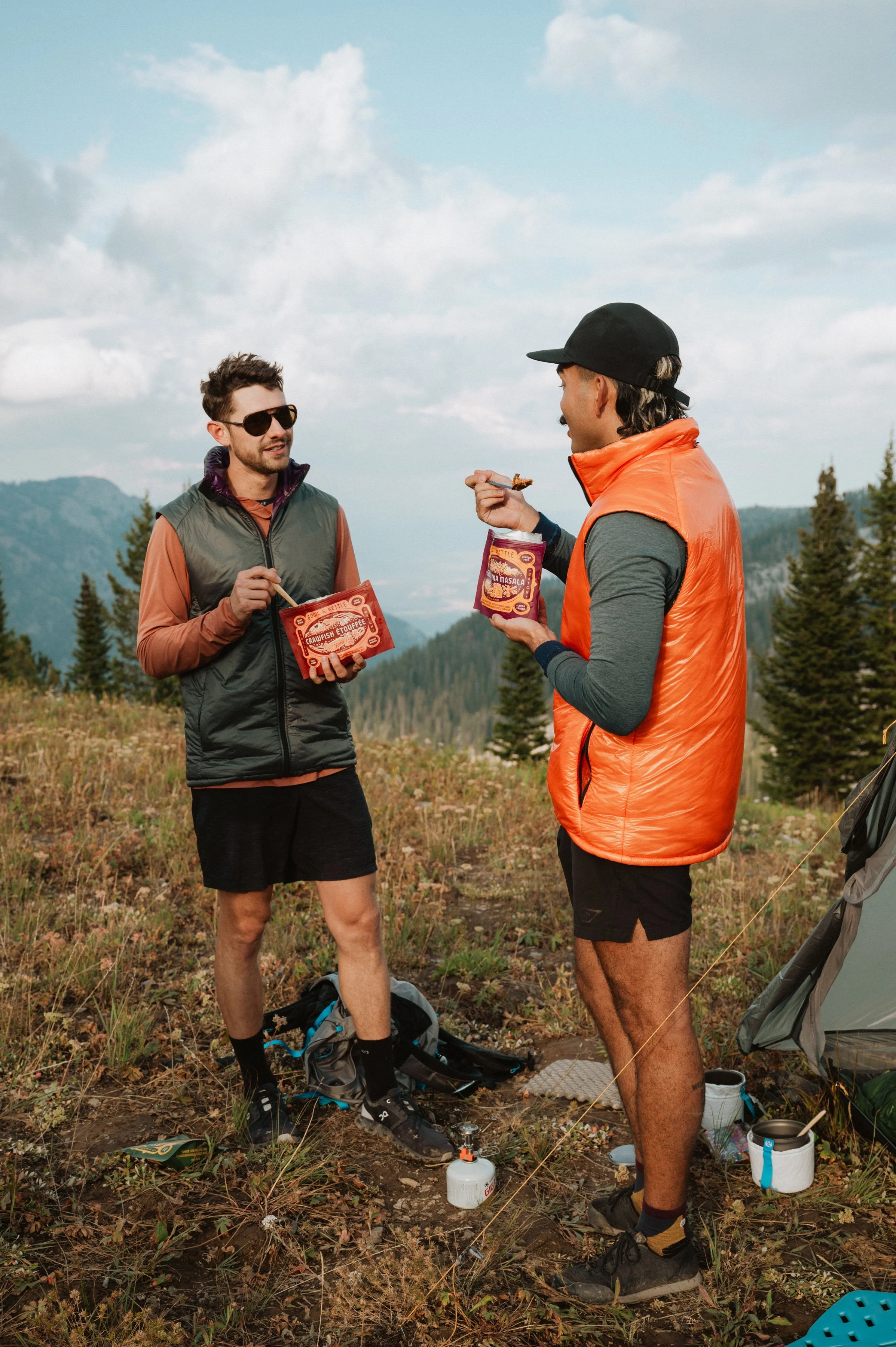 Two men camping outdoors on a mountain, talking and eating snacks, with a tent and camping gear around them.