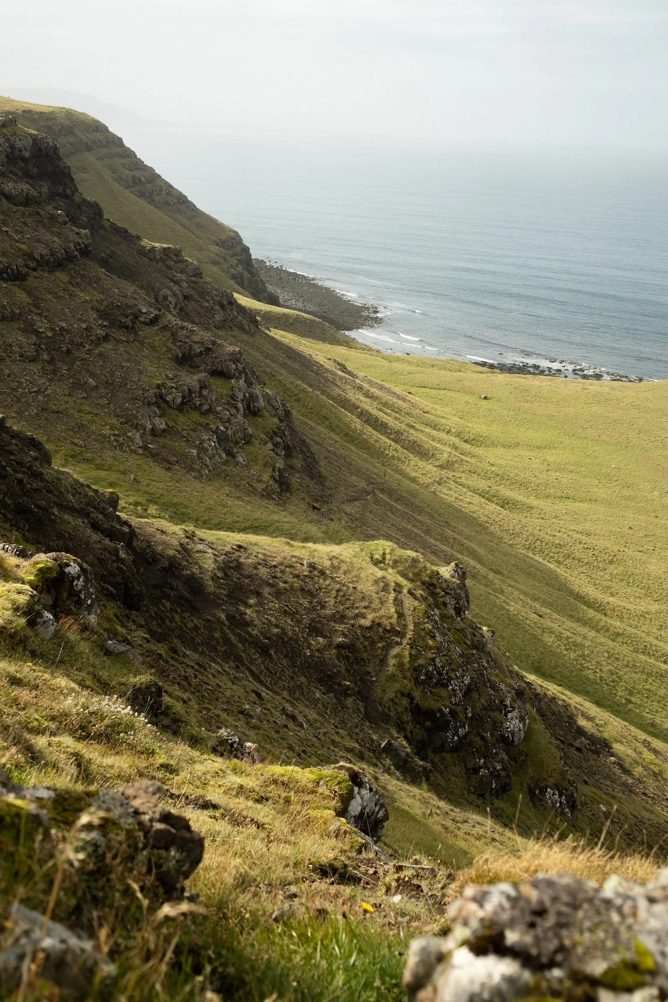 A scenic view of grassy, rocky hills leading down to the ocean, with a stretch of shoreline and the sea in the distance under cloudy skies.