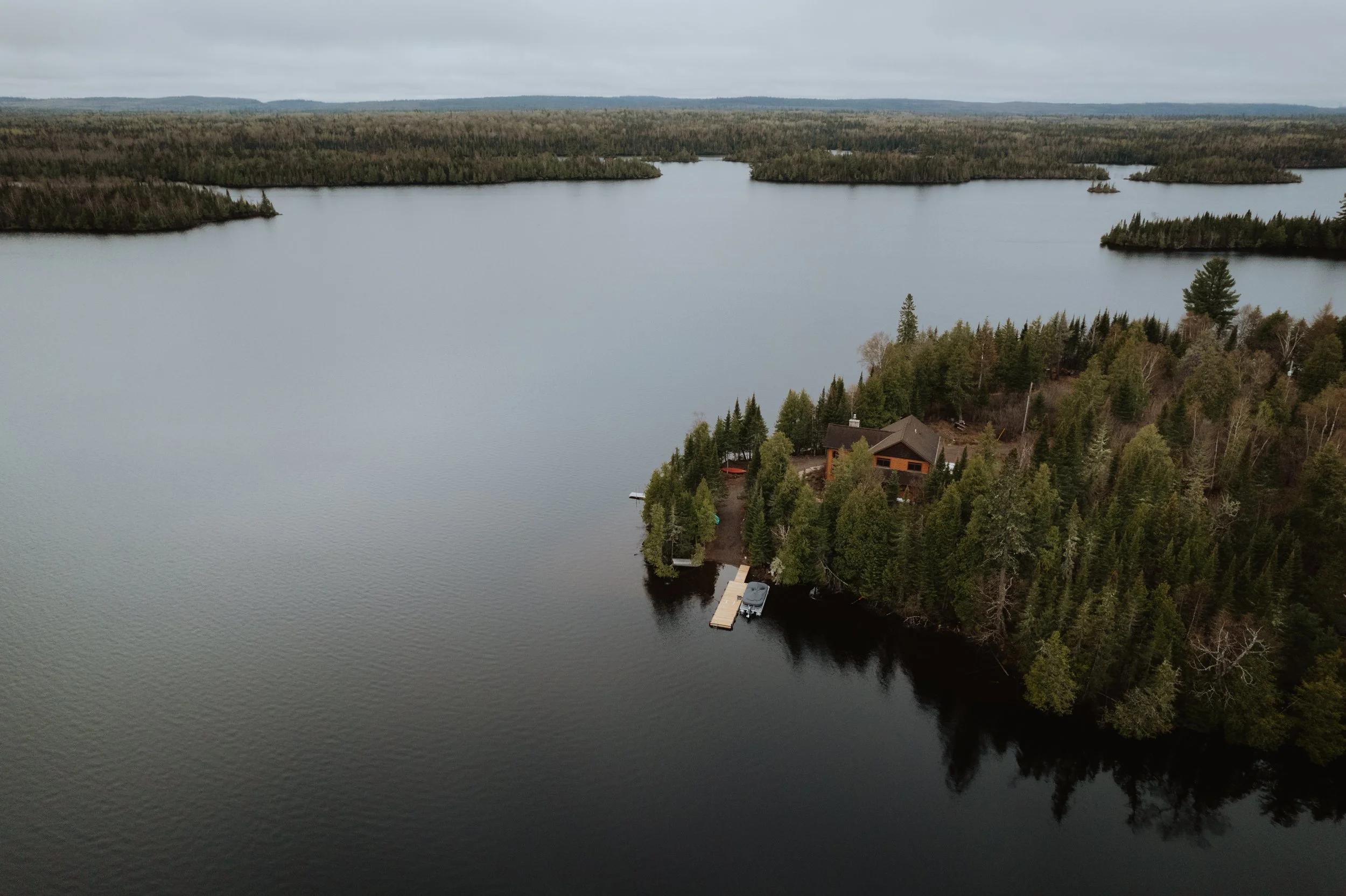 A house on a small forested island surrounded by a lake with a dock and boat, with a larger lake and forested land in the background.