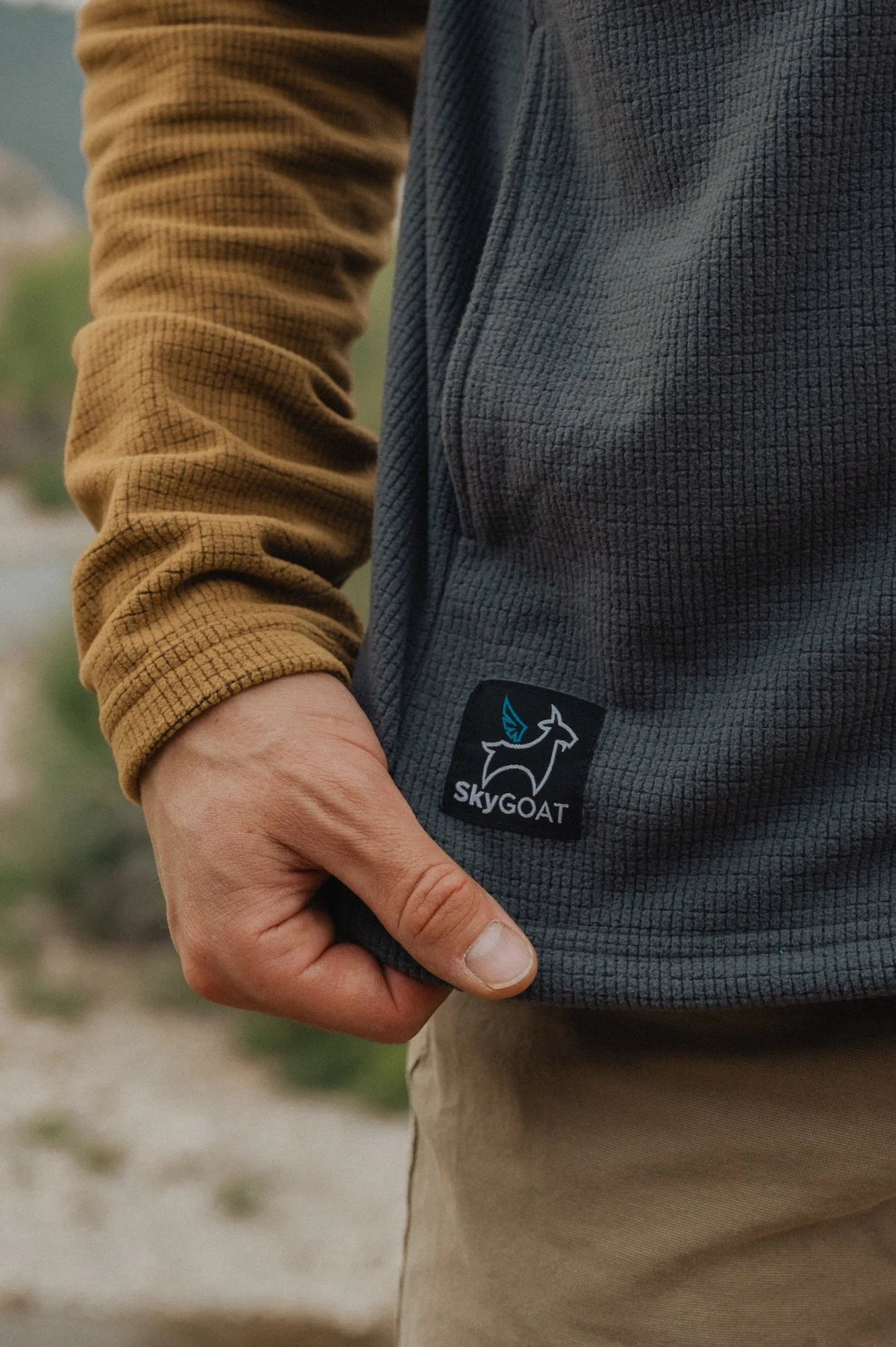 Close-up of a person wearing a gray fleece jacket with a SKYGOAT logo and a brown long-sleeve shirt underneath, with their hand in the jacket pocket, outdoors in a natural setting.