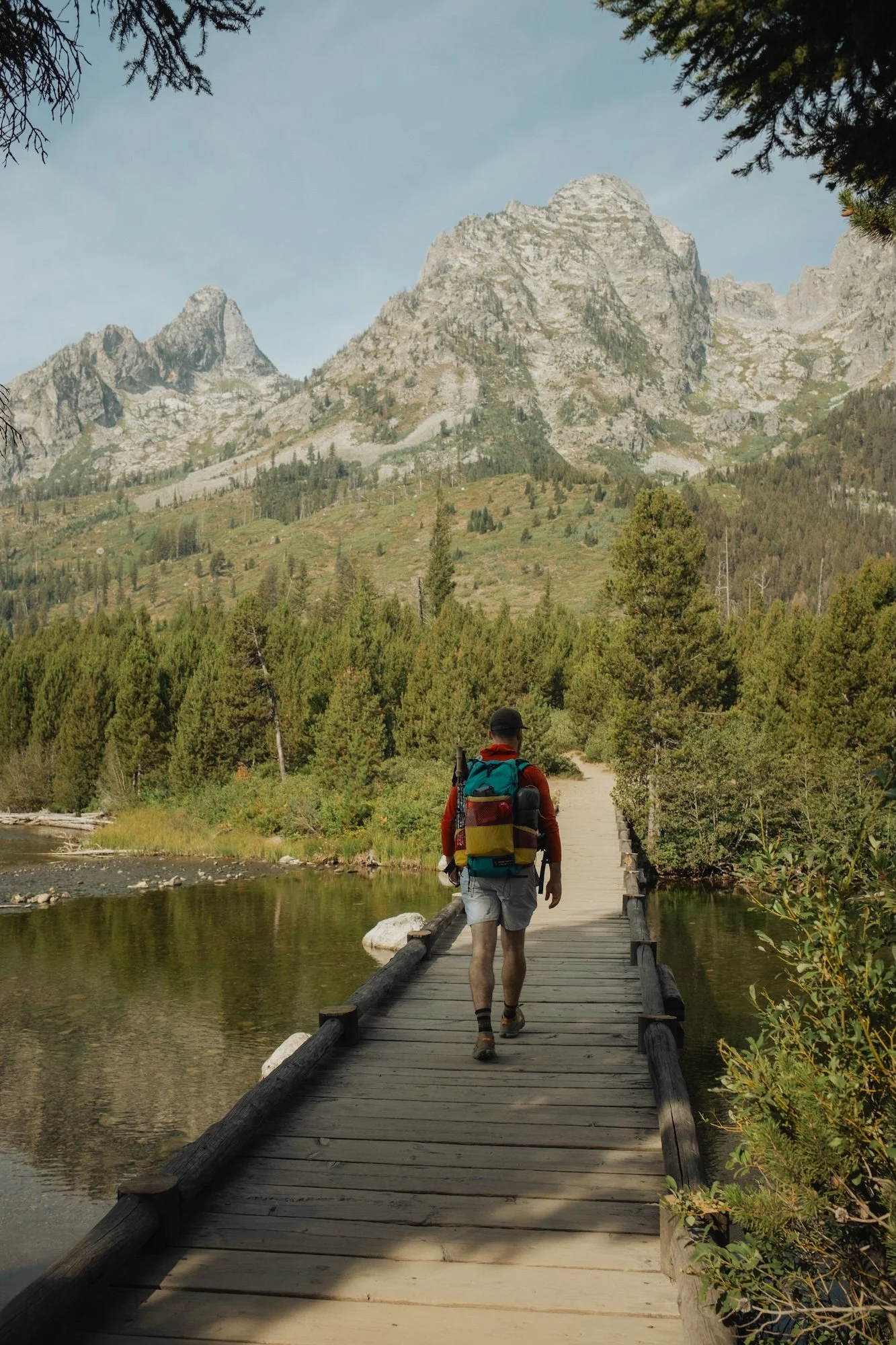 A person hiking across a wooden bridge over a river with mountains and trees in the background.