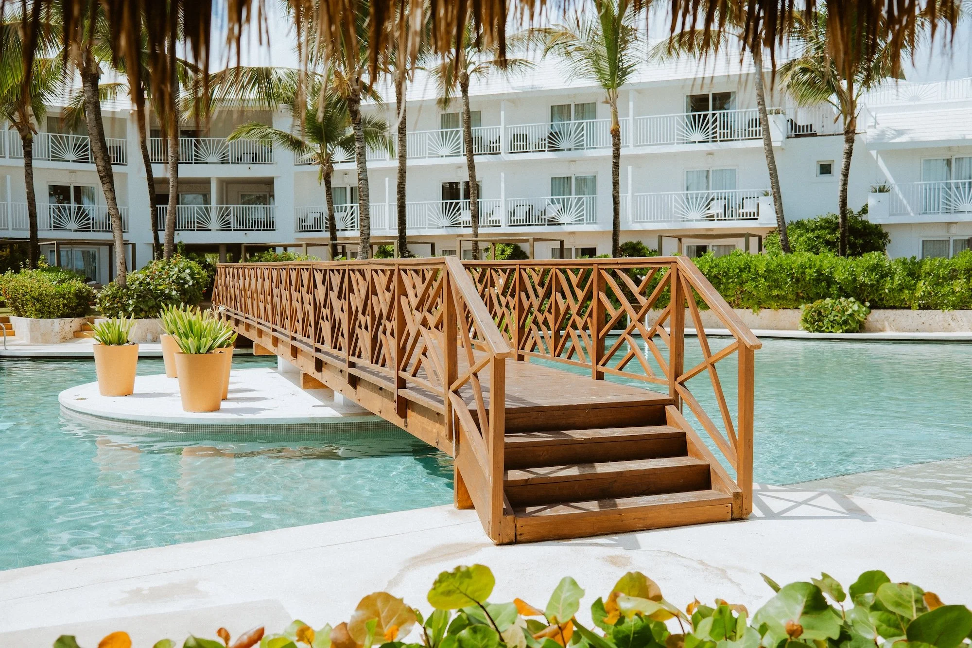 A small wooden bridge over a swimming pool at a resort, with potted plants and a white building with balconies in the background.