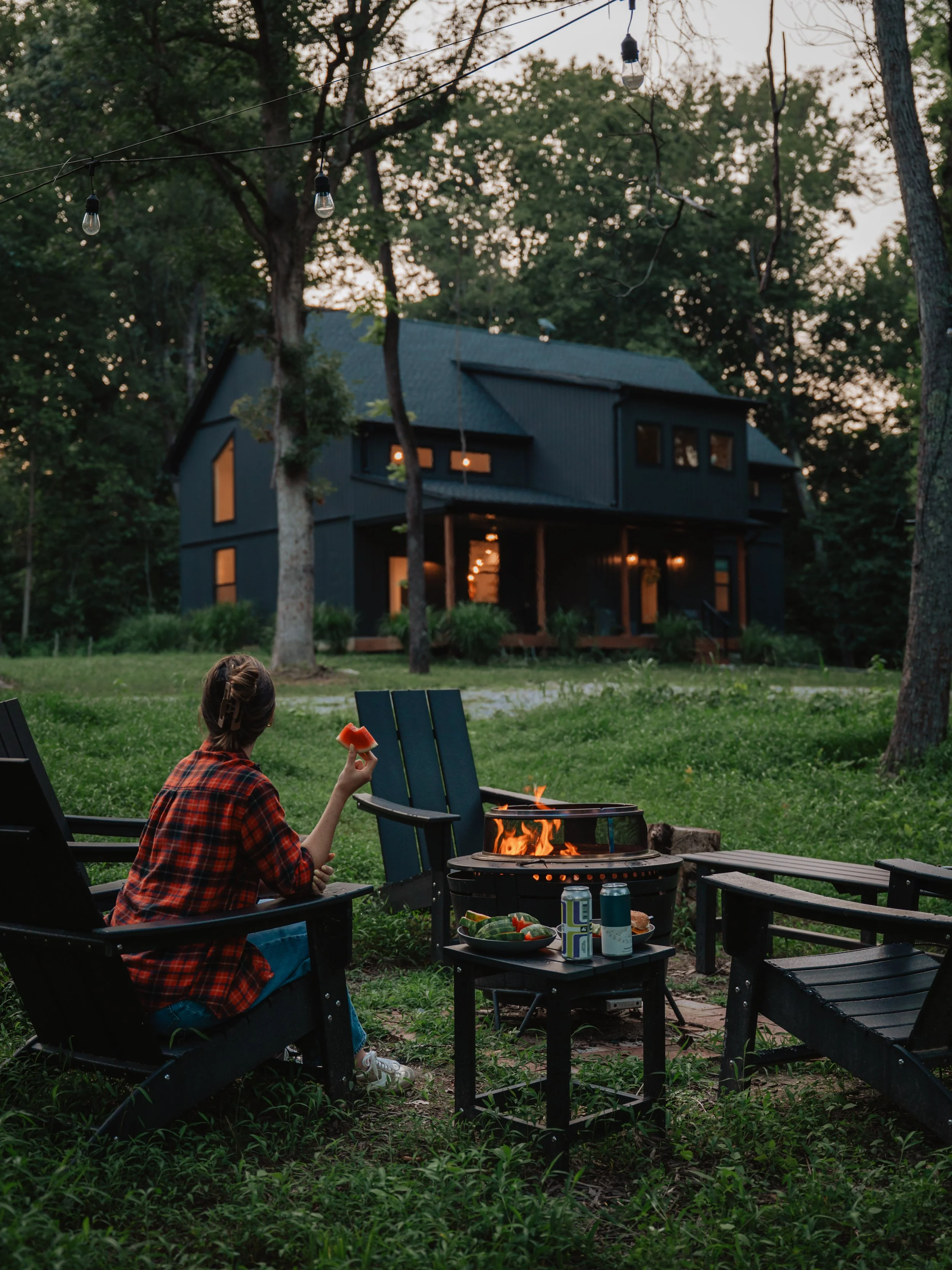 A person sitting in outdoor chairs near a fire pit grill, holding a slice of watermelon, with a modern black house and trees in the background at dusk.