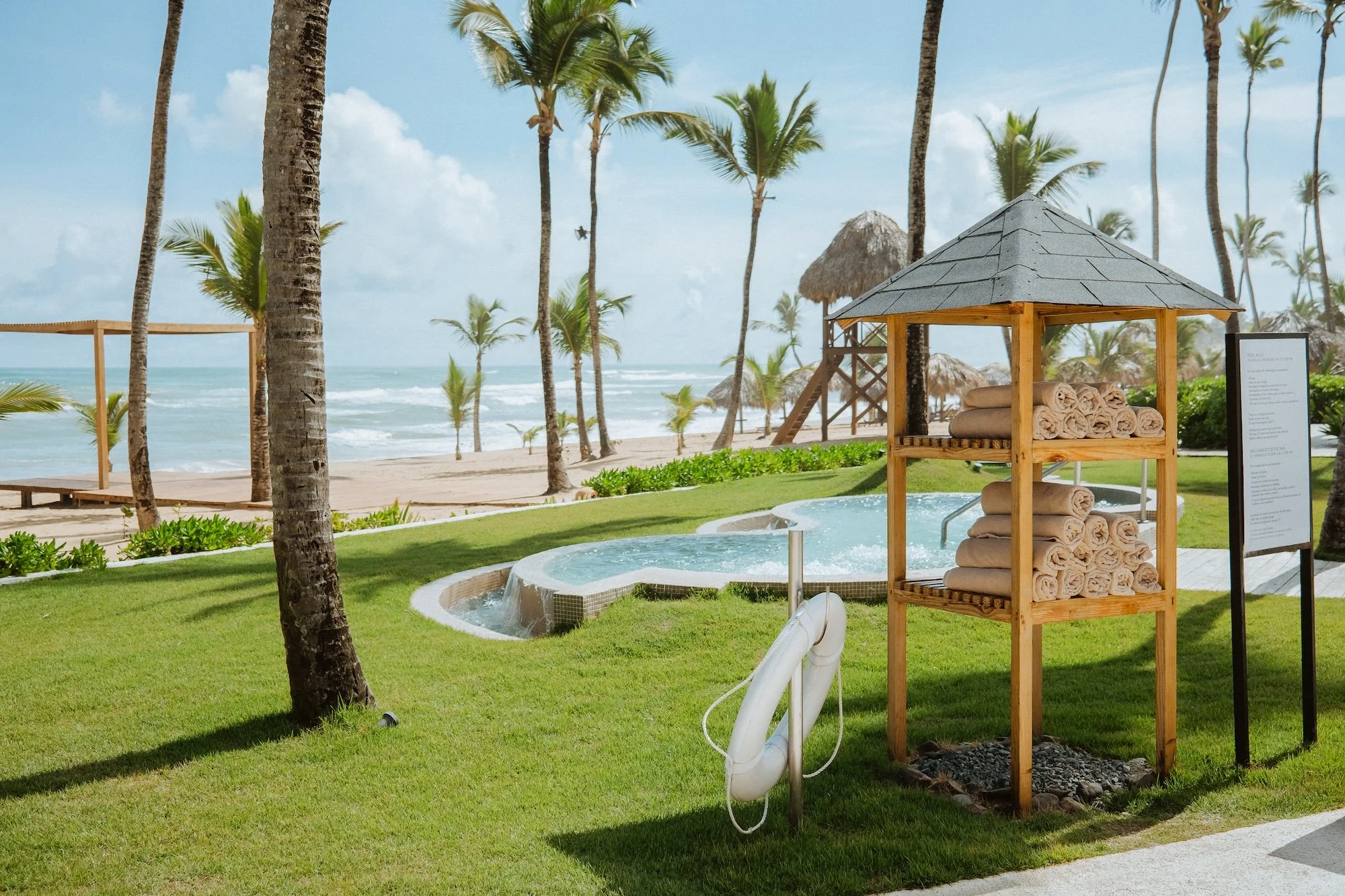 A tropical beach resort with palm trees, in-ground hot tub, with towels and a life preserver nearby, and sandy beach with ocean waves in the background