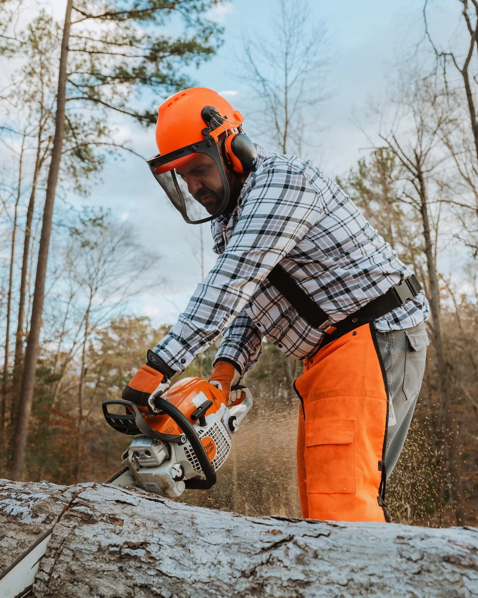 Man cutting a large fallen tree log with a chainsaw in a wooded outdoor area during daytime.