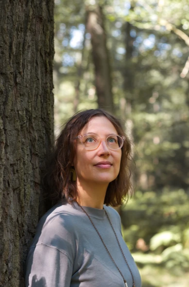 Frau mit rosa Brille lehnt gegen einen Baumstamm und schaut glücklich lächelnd nach oben