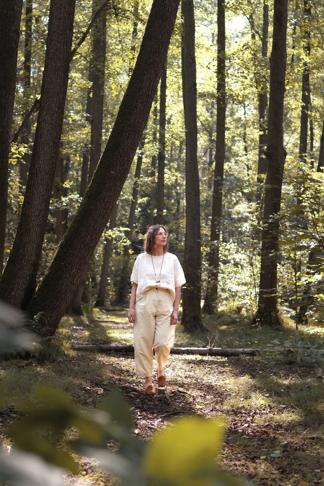 Eine Frau in hellen Kleidung spaziert im Wald auf einem Pfad, umgeben von Bäumen und grünen Blättern, bei Sonnenlicht.