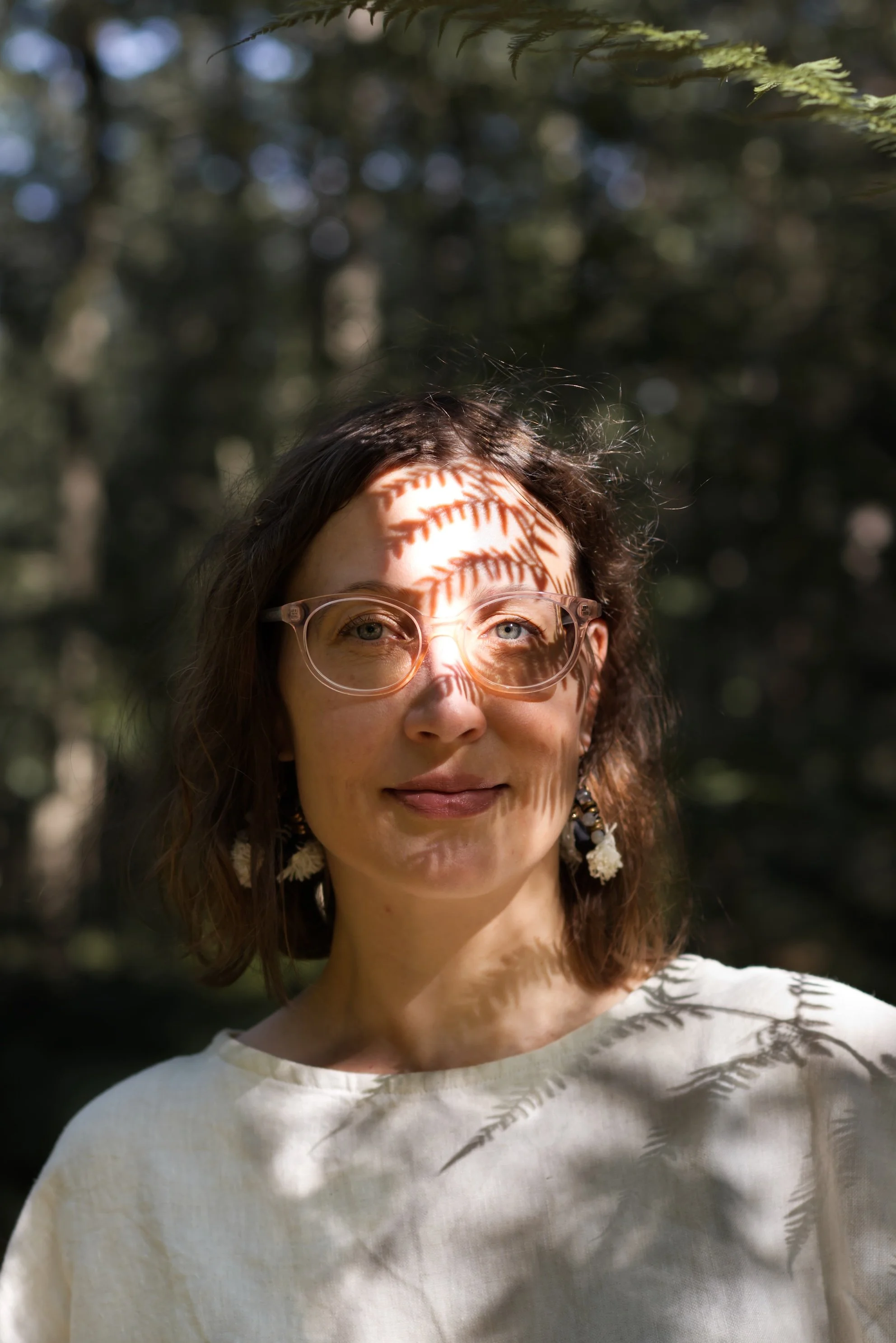 Frau in hellem Oberteil vor dunklem Wald und mit Schattenmuster eines Farns über dem Gesicht
