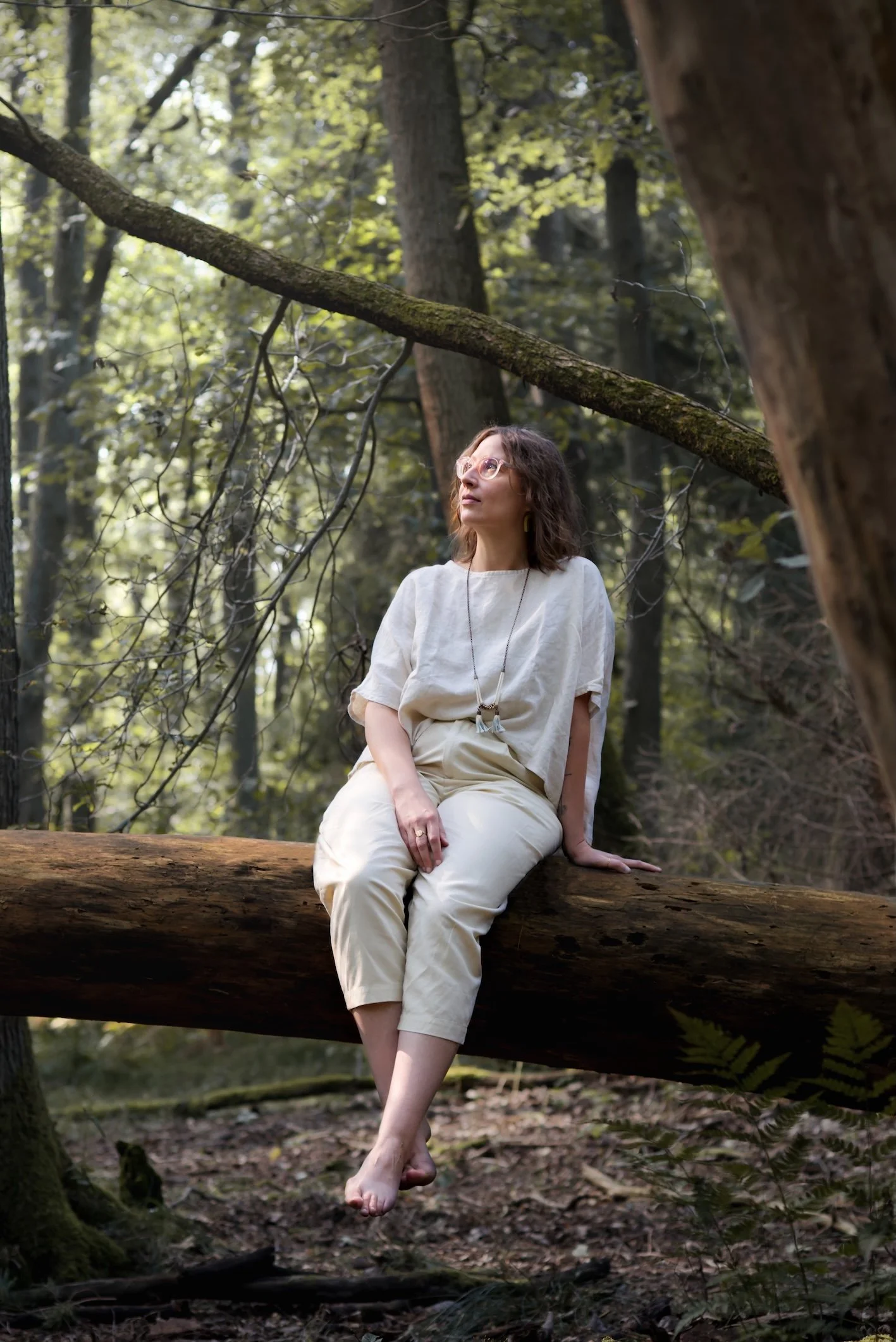 Frau in heller Leinenkleidung sitzt entspannt auf einem waagrechten Baumstamm