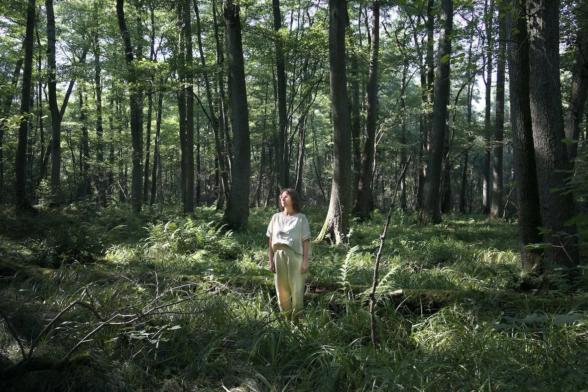 Eine Frau steht in einem dichten grünen Wald, umgeben von hohen Bäumen und üppigem Unterholz, während Sonnenlicht durch die Baumkronen fällt.