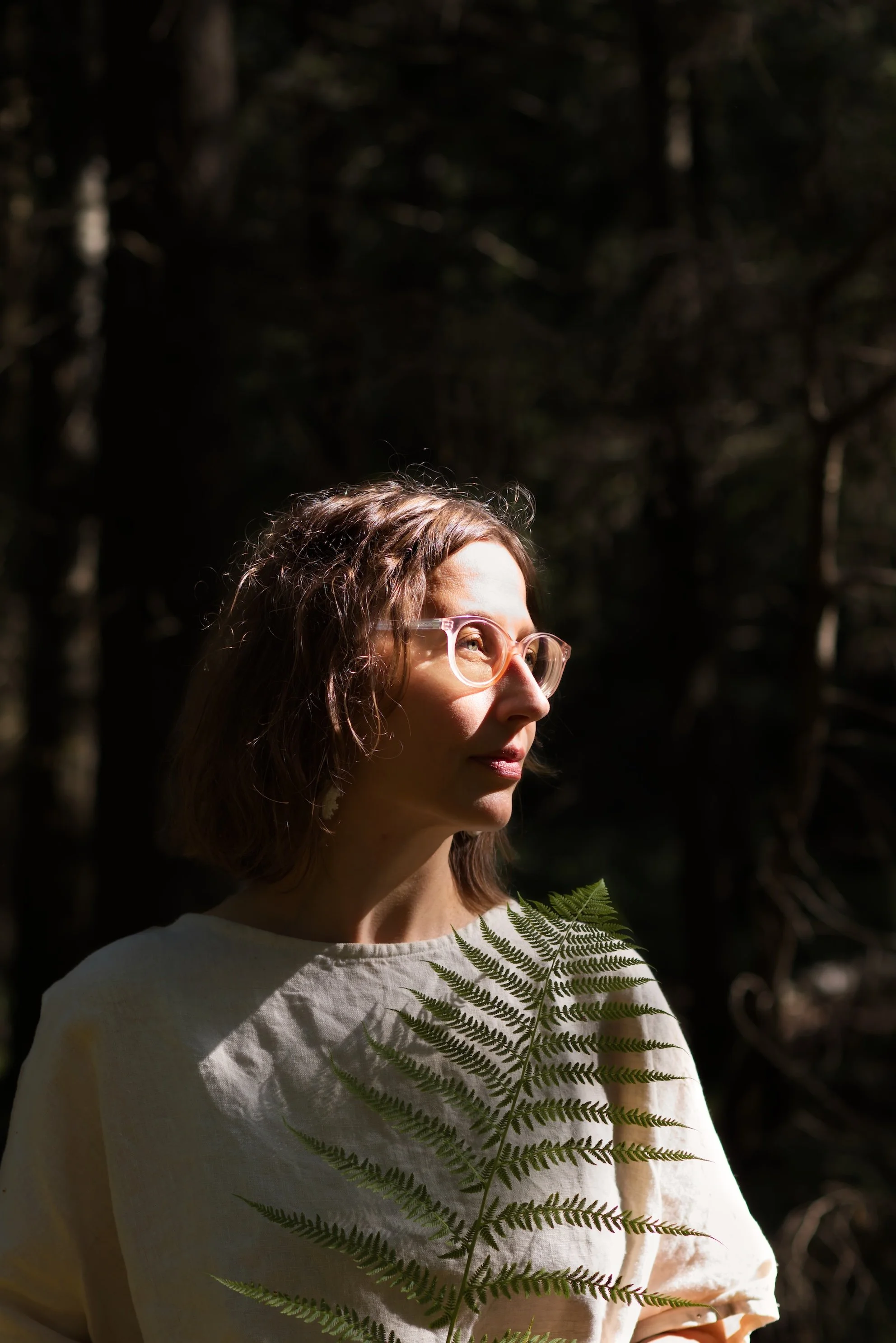 Frau mit rosa Brille, hellem Shirt und Farn vor dem Oberkörper wird von Sonnenlicht beleuchtet
