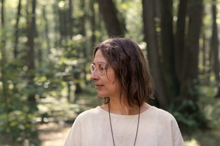 Frau in einer hellen Leinenbluse steht im Wald und lauscht in die Stille