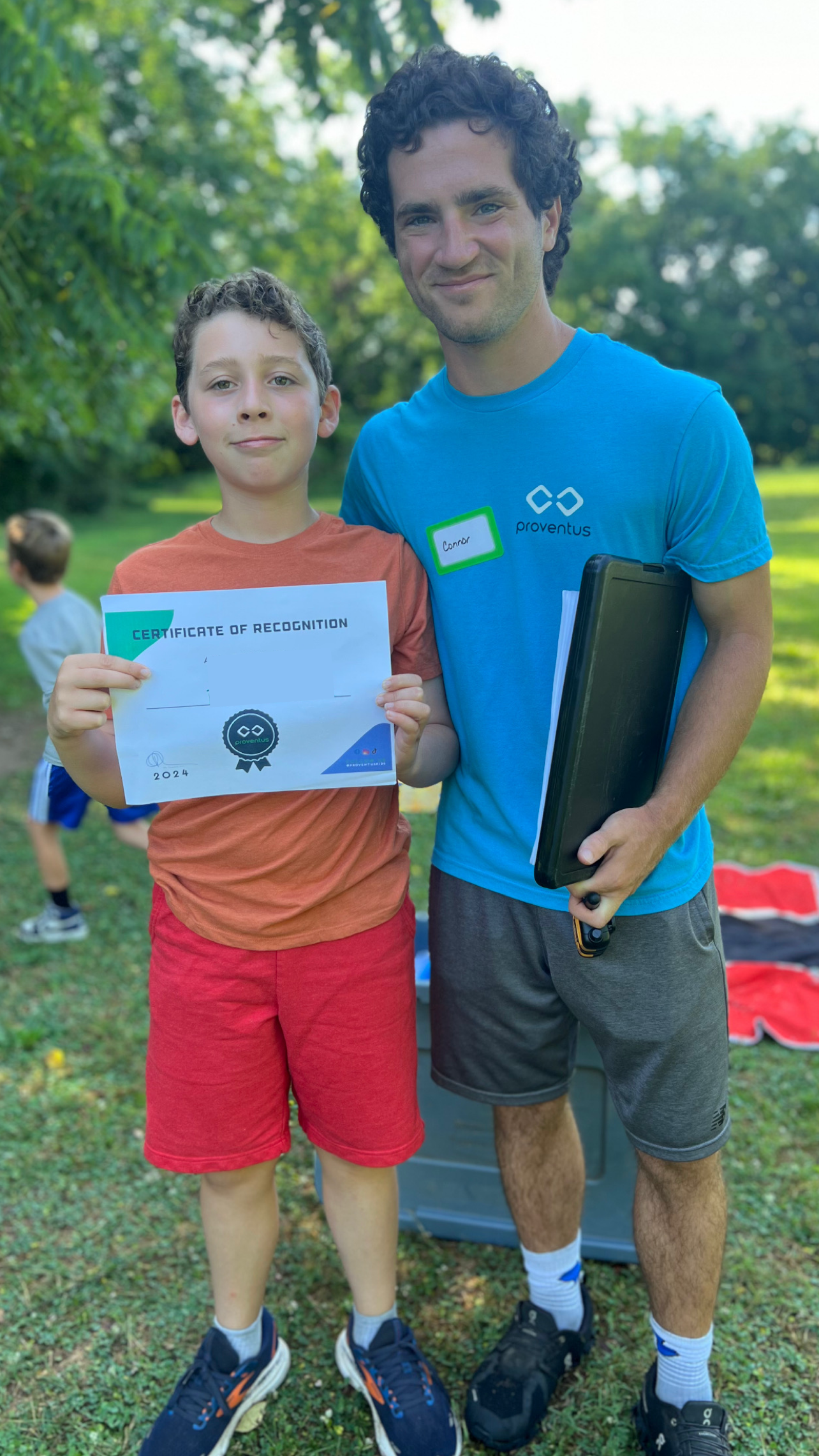 A young boy holding a certificate of recognition while standing next to a man in a blue shirt, outdoors in a park with children in the background.
