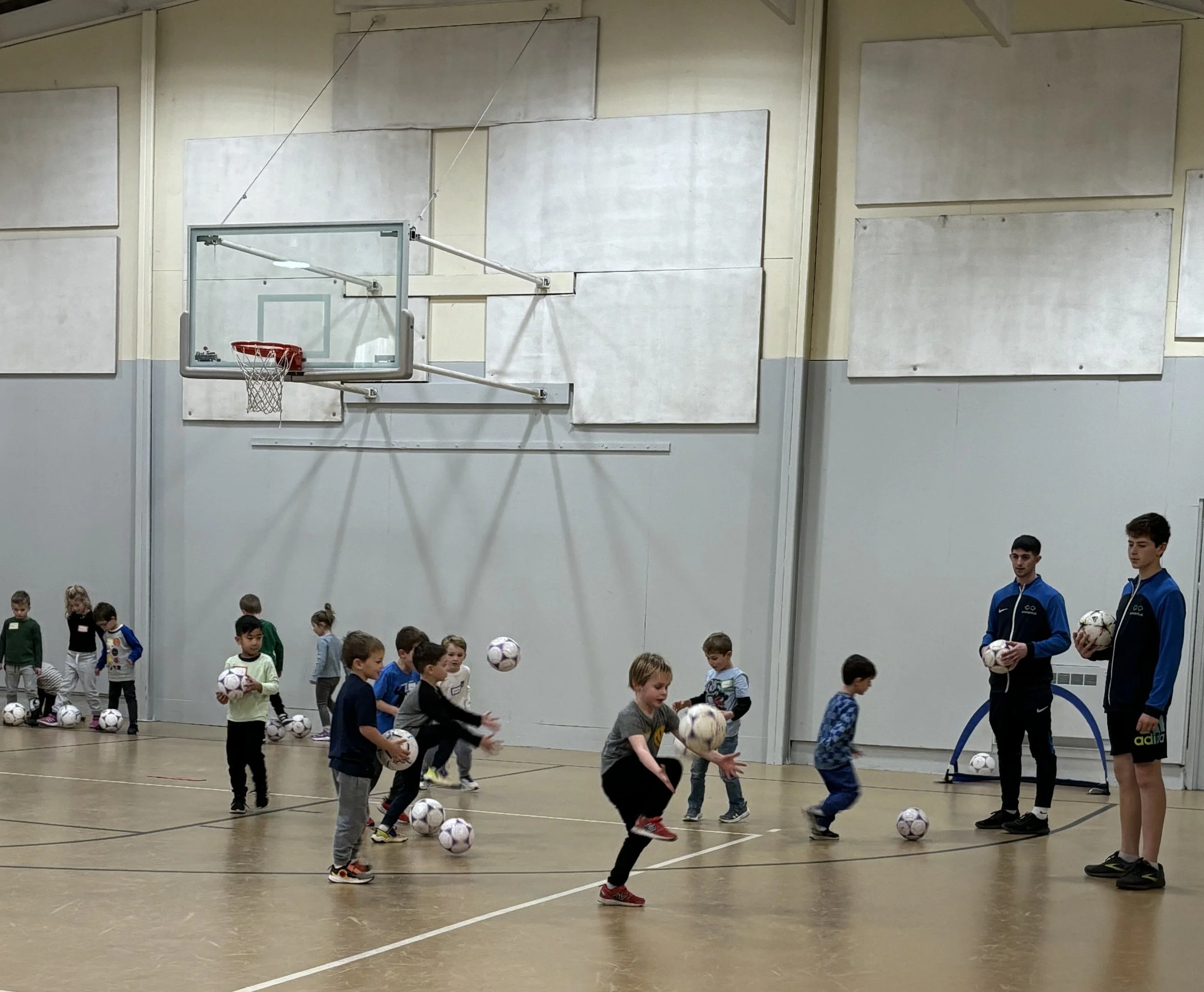 Group of children participating in a soccer training session indoors, with two coaches holding soccer balls. Children are dribbling and passing soccer balls on the basketball court.