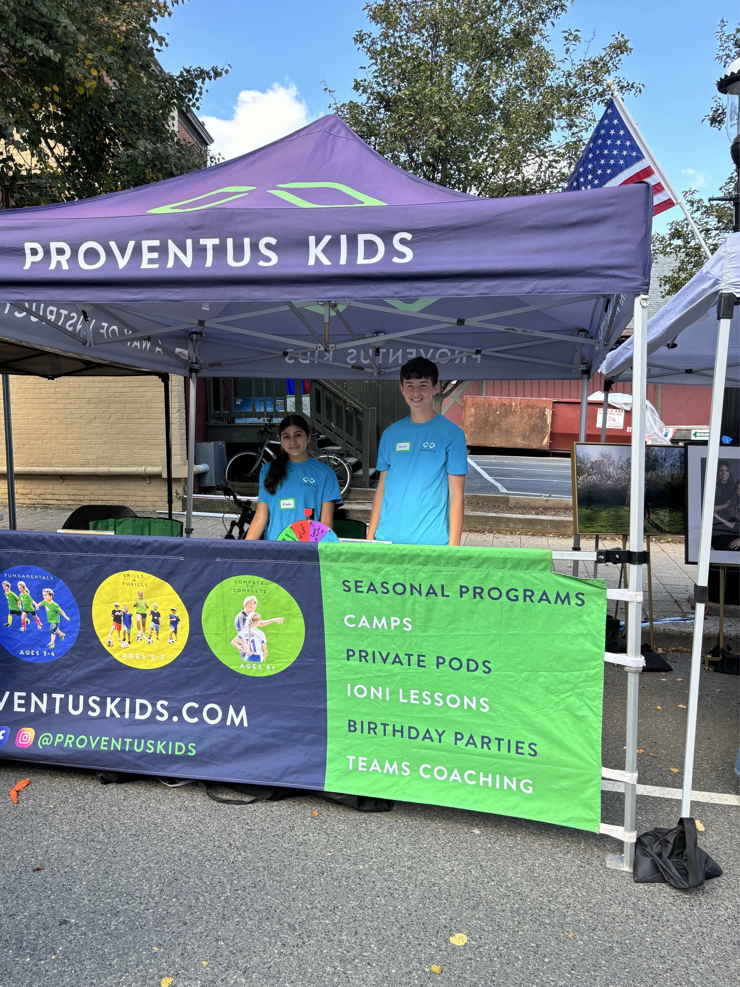 A booth for Proventus Kids with two young volunteers standing behind a table at an outdoor event, under a purple canopy with the logo, and a green banner listing their programs such as camps, private pods, Ioni lessons, birthday parties, and teams coaching.
