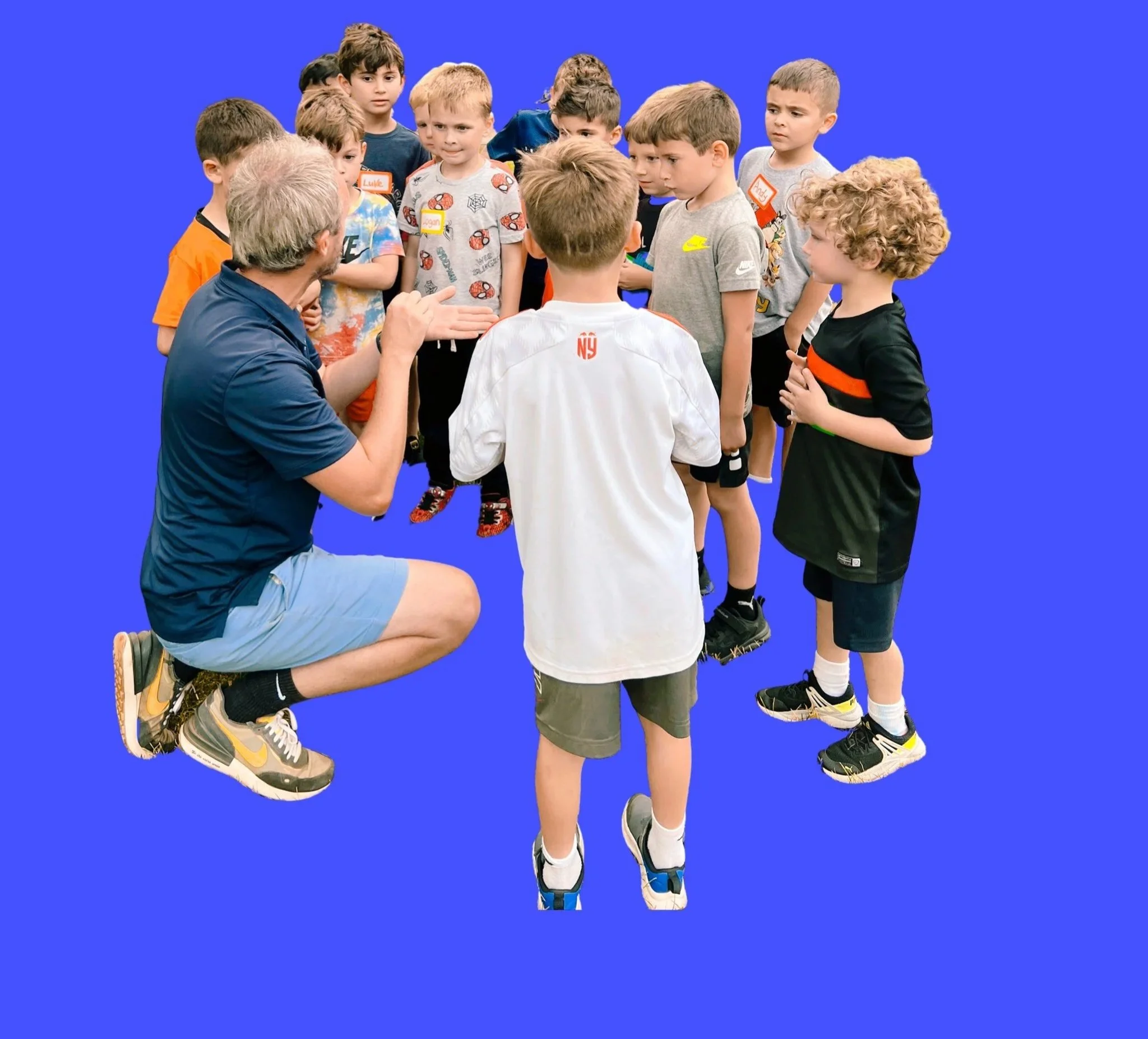 A group of young boys in casual sportswear standing in a semi-circle around a man who is kneeling and talking to them. The background is bright blue.
