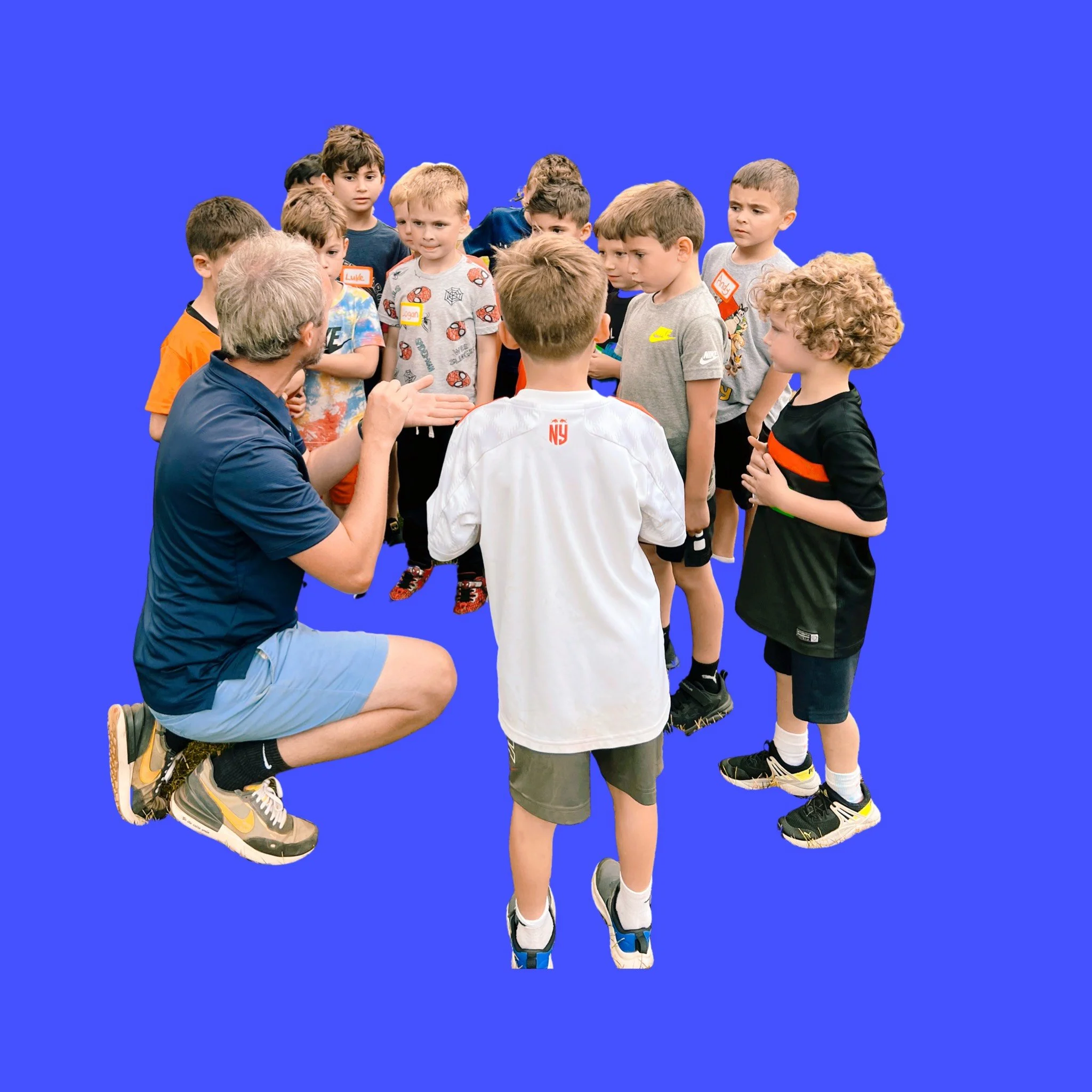 A group of young boys standing together, one kneeling, with a man talking to them against a solid blue background.