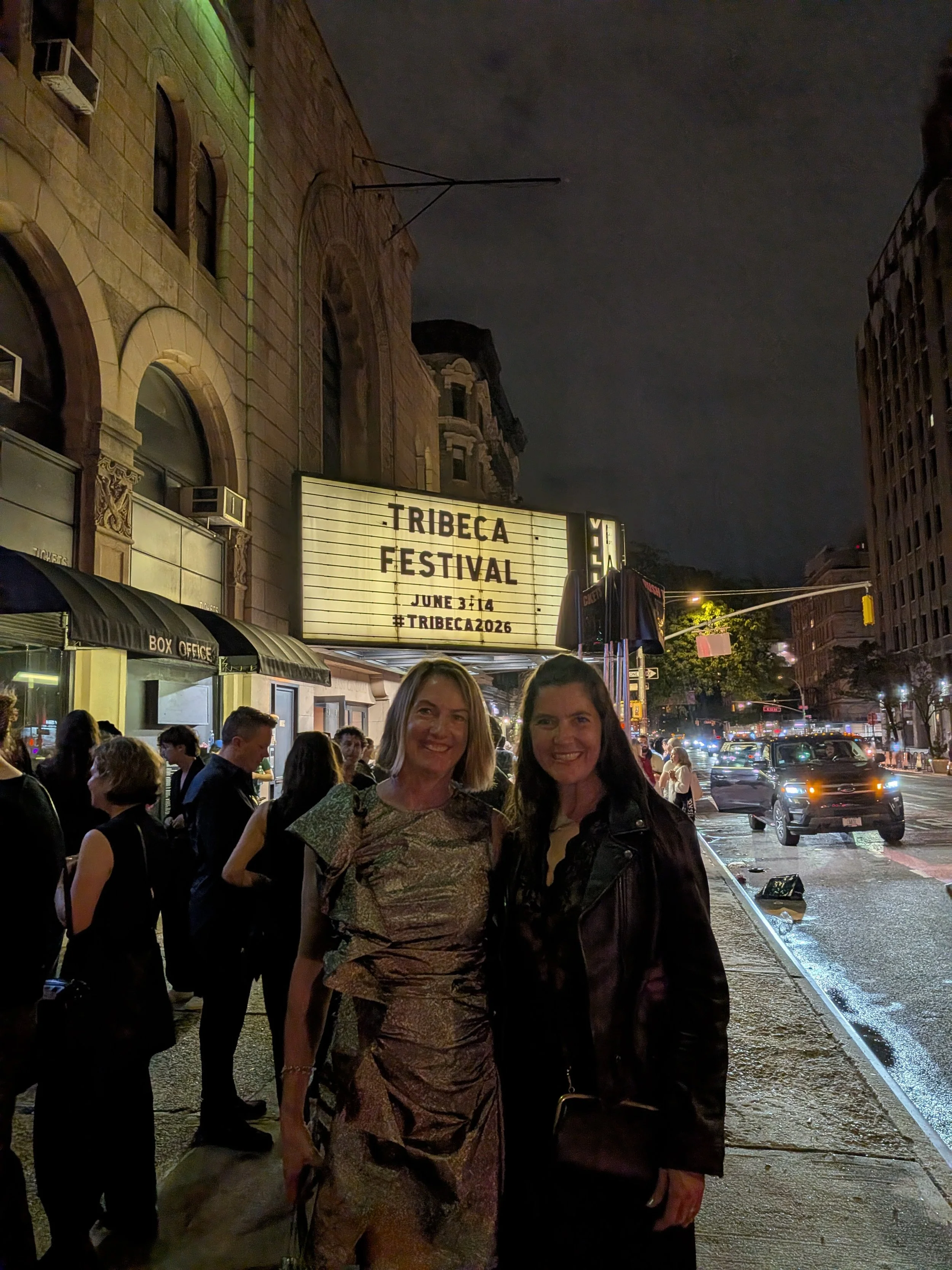 Nighttime street scene with a crowd of people, two women posing for a photo in the foreground, a marquee sign reading 'Tribeca Festival June 3-14 #Tribeca2026', and cars on a wet street.