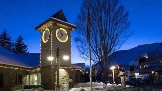 A clock tower decorated with Christmas lights outdoors at dusk or early evening, with snow on the ground and nearby buildings.