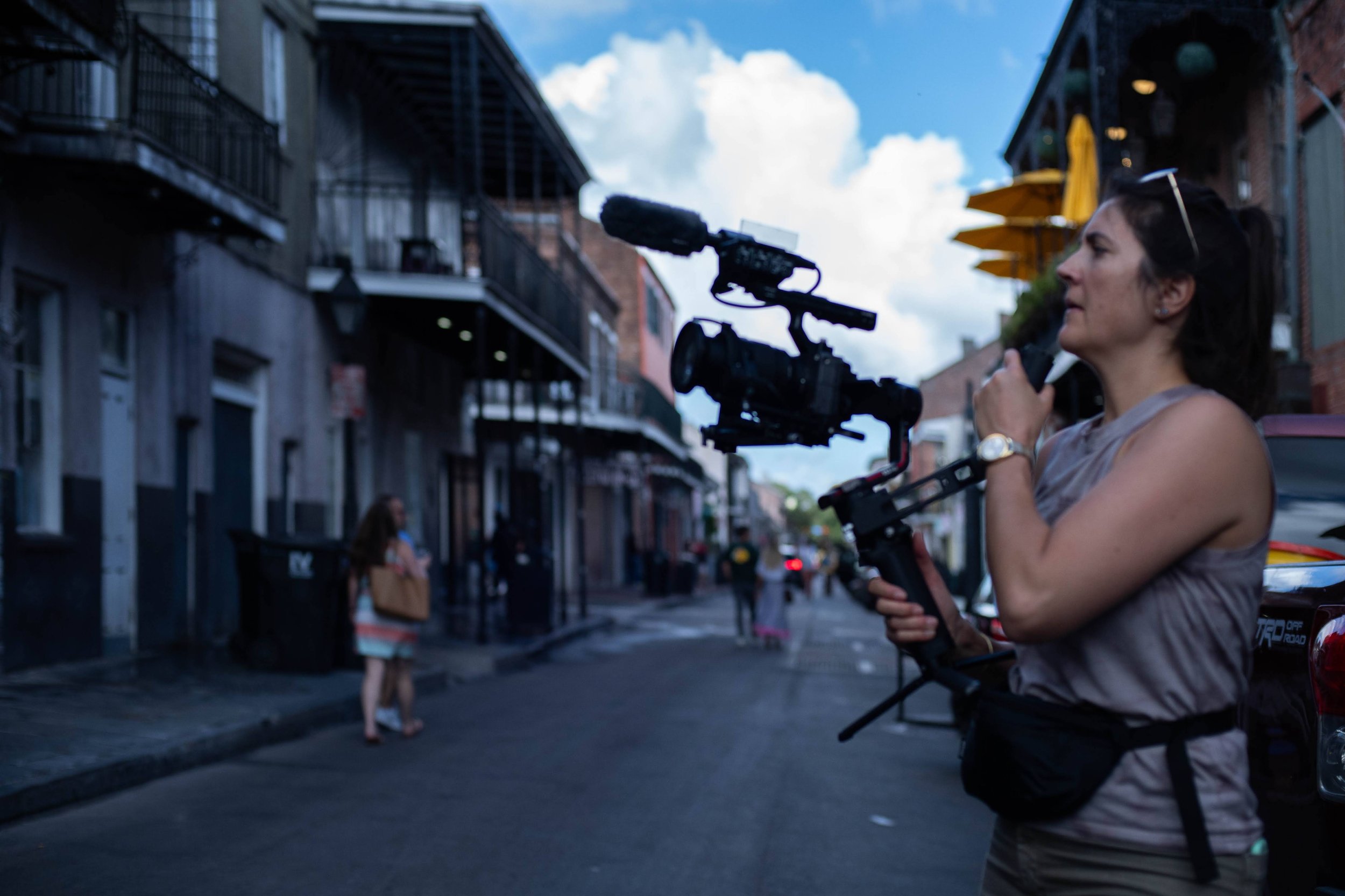 A woman operating a professional video camera on a street in an urban setting, with buildings and pedestrians in the background during daytime.