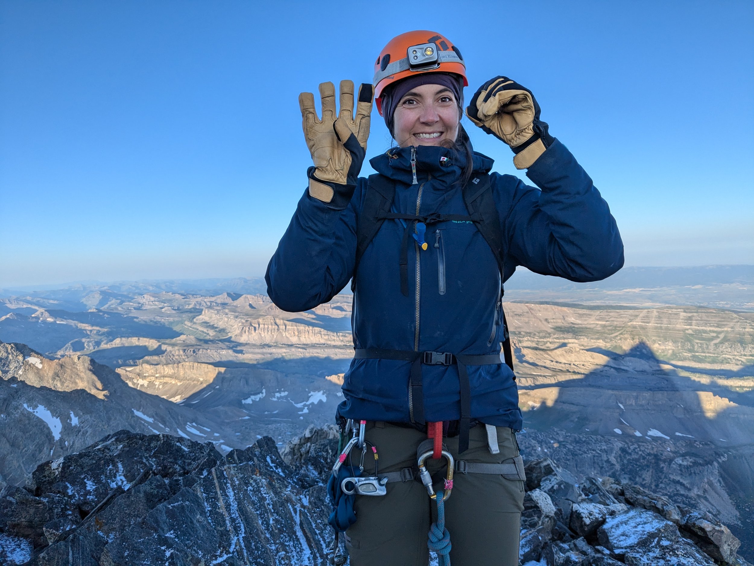 A woman in mountain climbing gear, wearing a helmet and smiling, standing on a rocky mountain summit with snow patches and a vast mountainous landscape in the background.