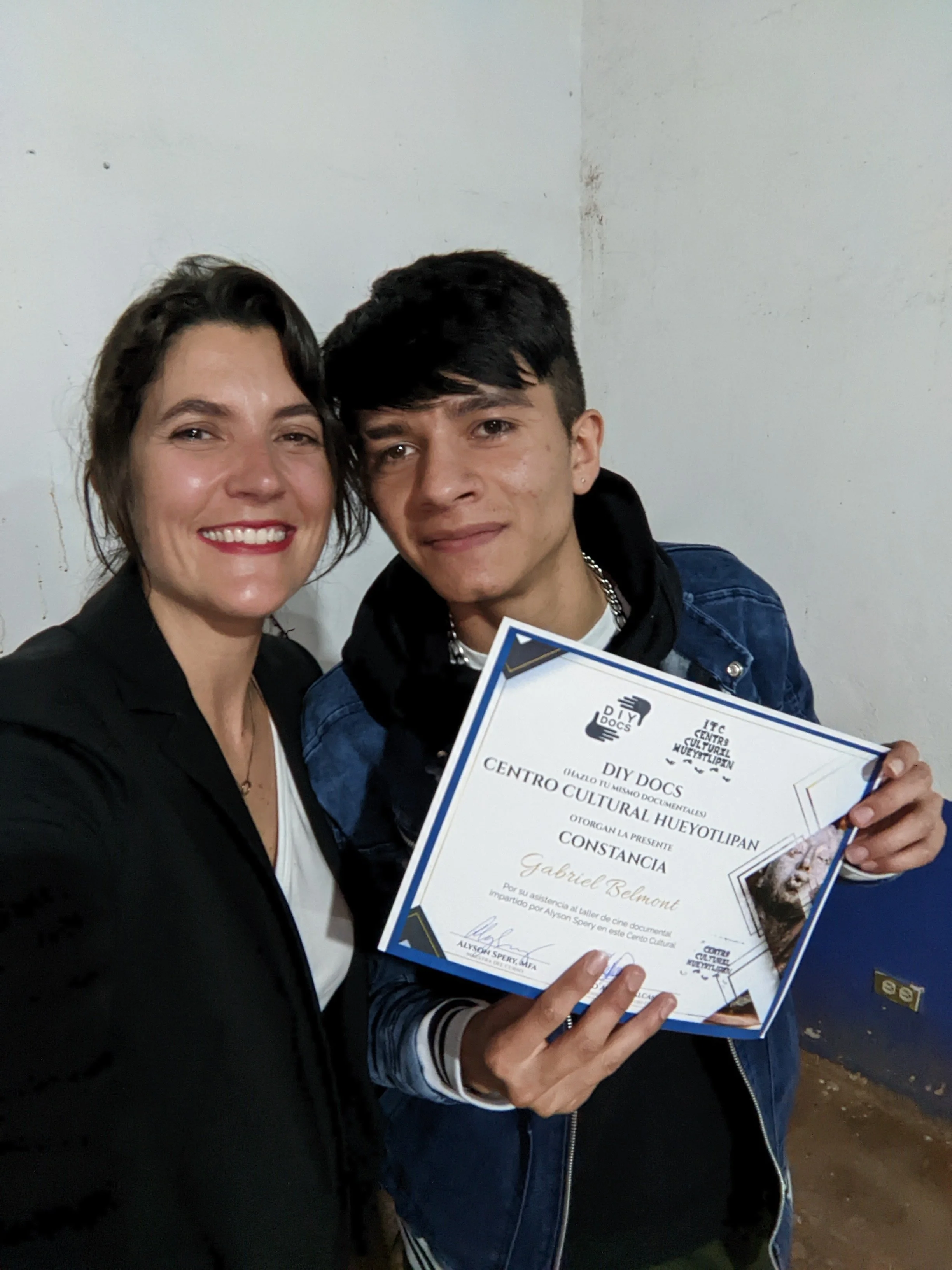 Two people, a woman with short dark hair and a young man with short black hair, are smiling and holding a certificate together. They are standing indoors against a plain wall.