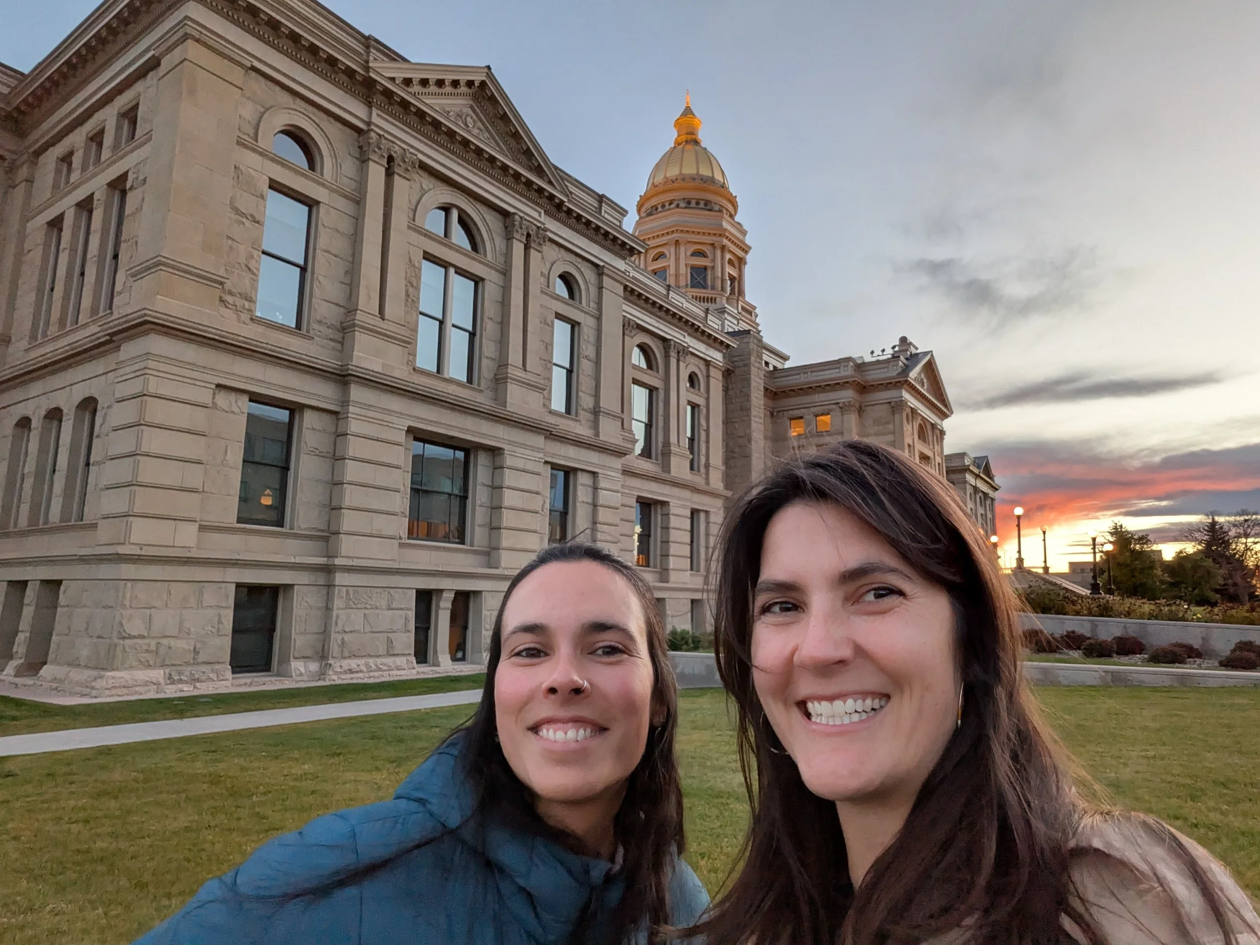 Two smiling women taking a selfie in front of a historic building with large windows, stone architecture, and a dome, during sunset.