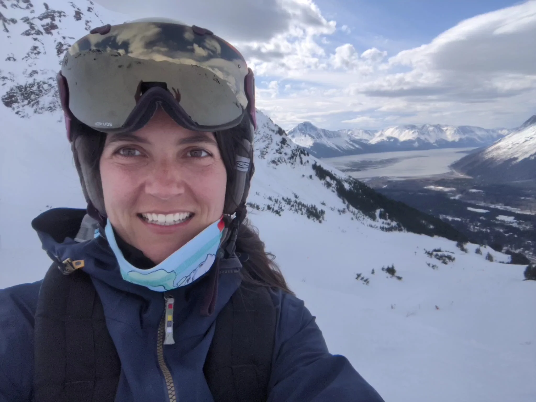A woman wearing a ski helmet and goggles smiling on snowy mountains with a partly cloudy sky in the background.