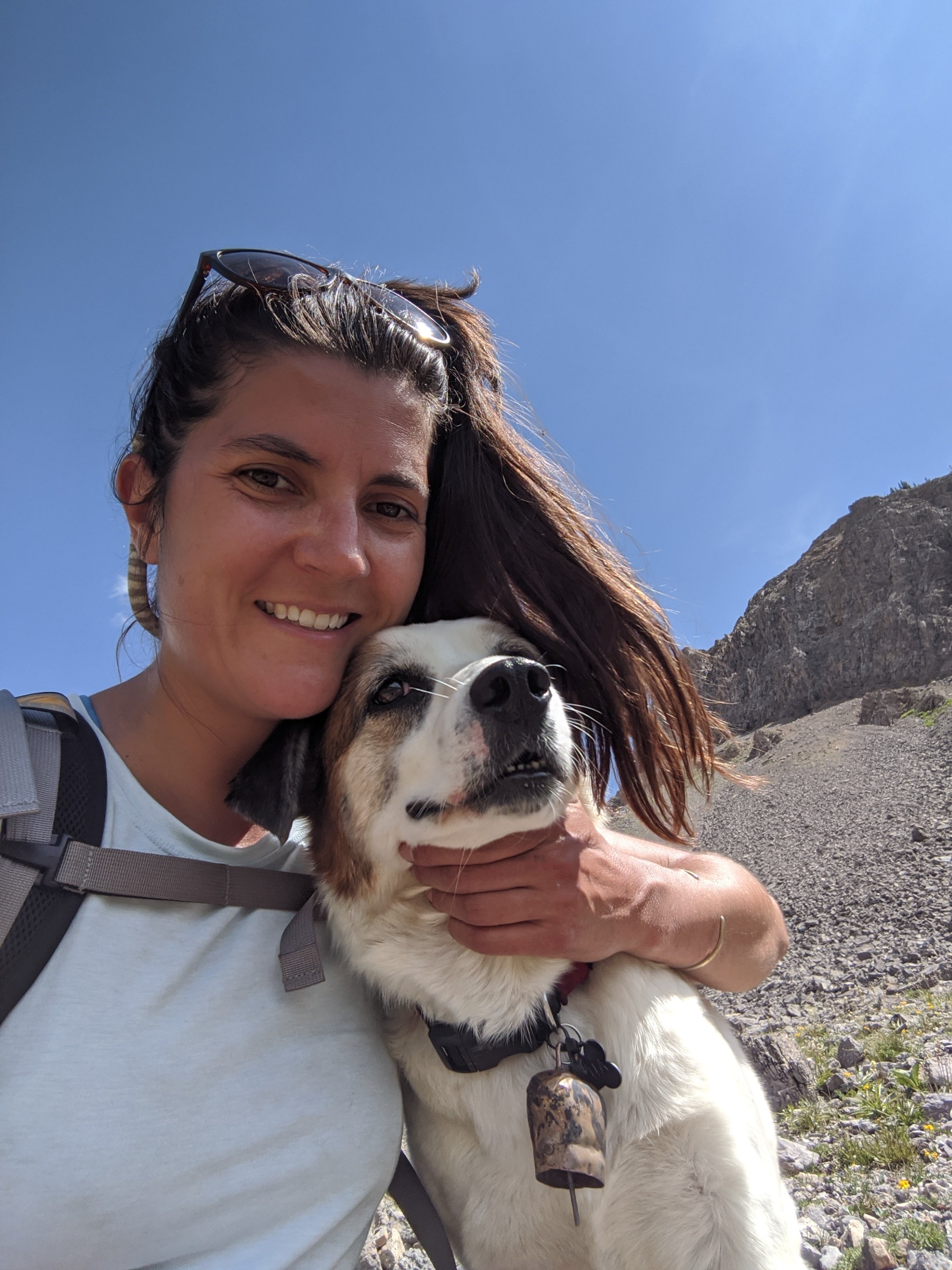 A woman with sunglasses on her head, smiling, holding a large white and brown dog with a black collar. They are outside on a rocky terrain with mountains and a clear blue sky in the background.