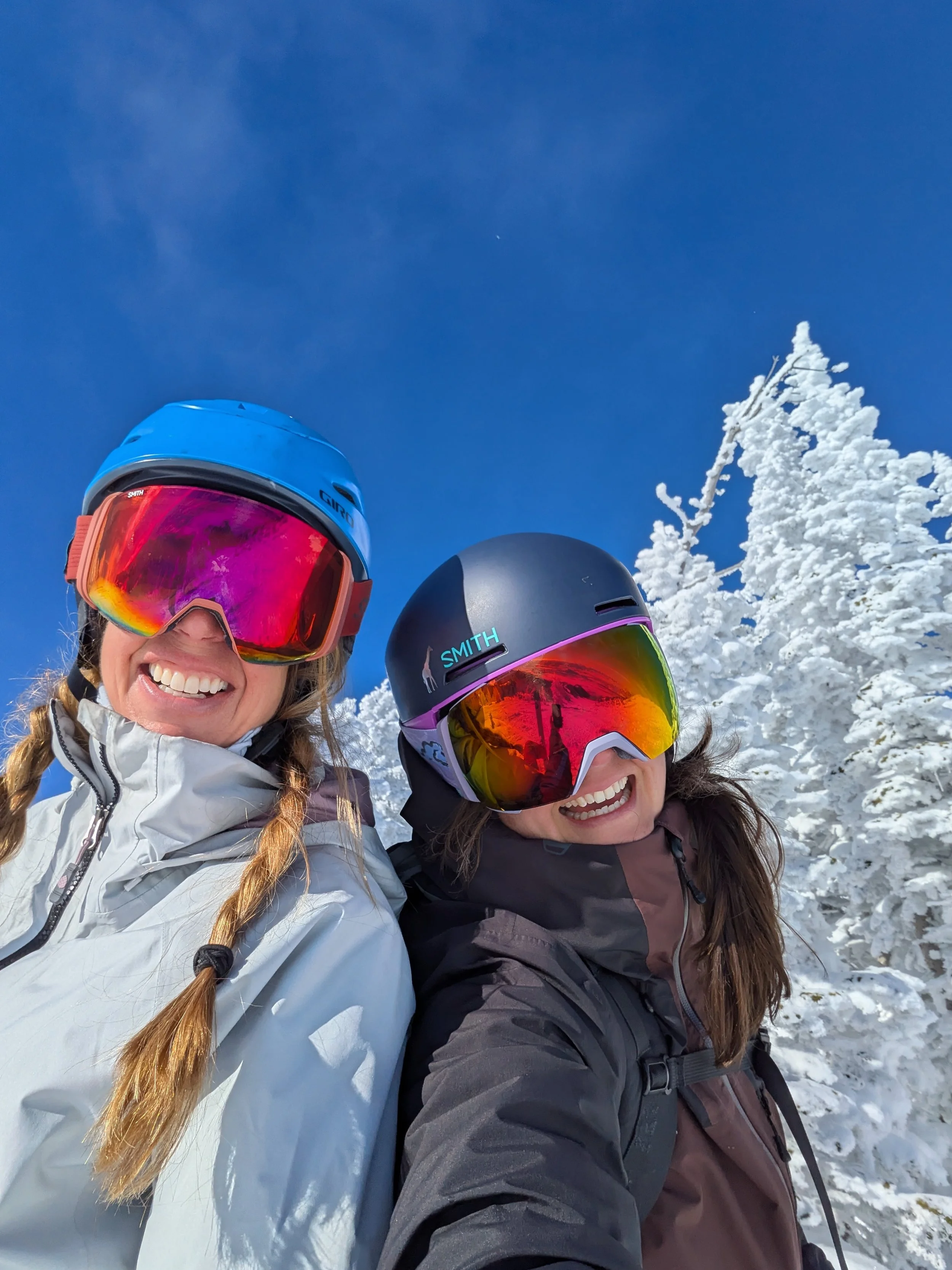 Two women in winter clothing, wearing ski helmets and goggles, smiling for a selfie with snow-covered trees and a clear blue sky in the background.