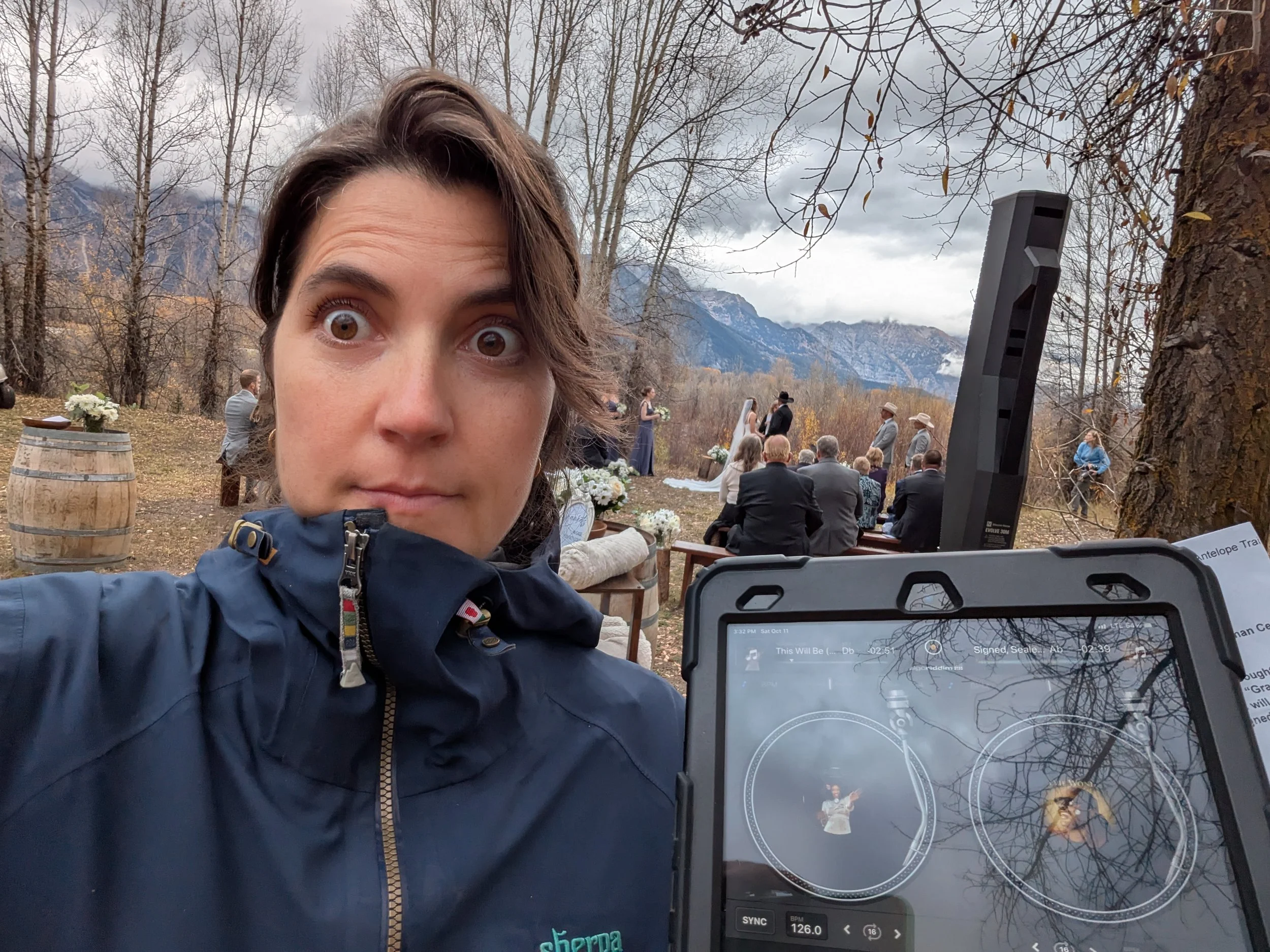 A woman taking a selfie outdoors during a cloudy day at a mountain wedding venue with seated guests, a wedding arch, trees with fall leaves, and mountains in the background.