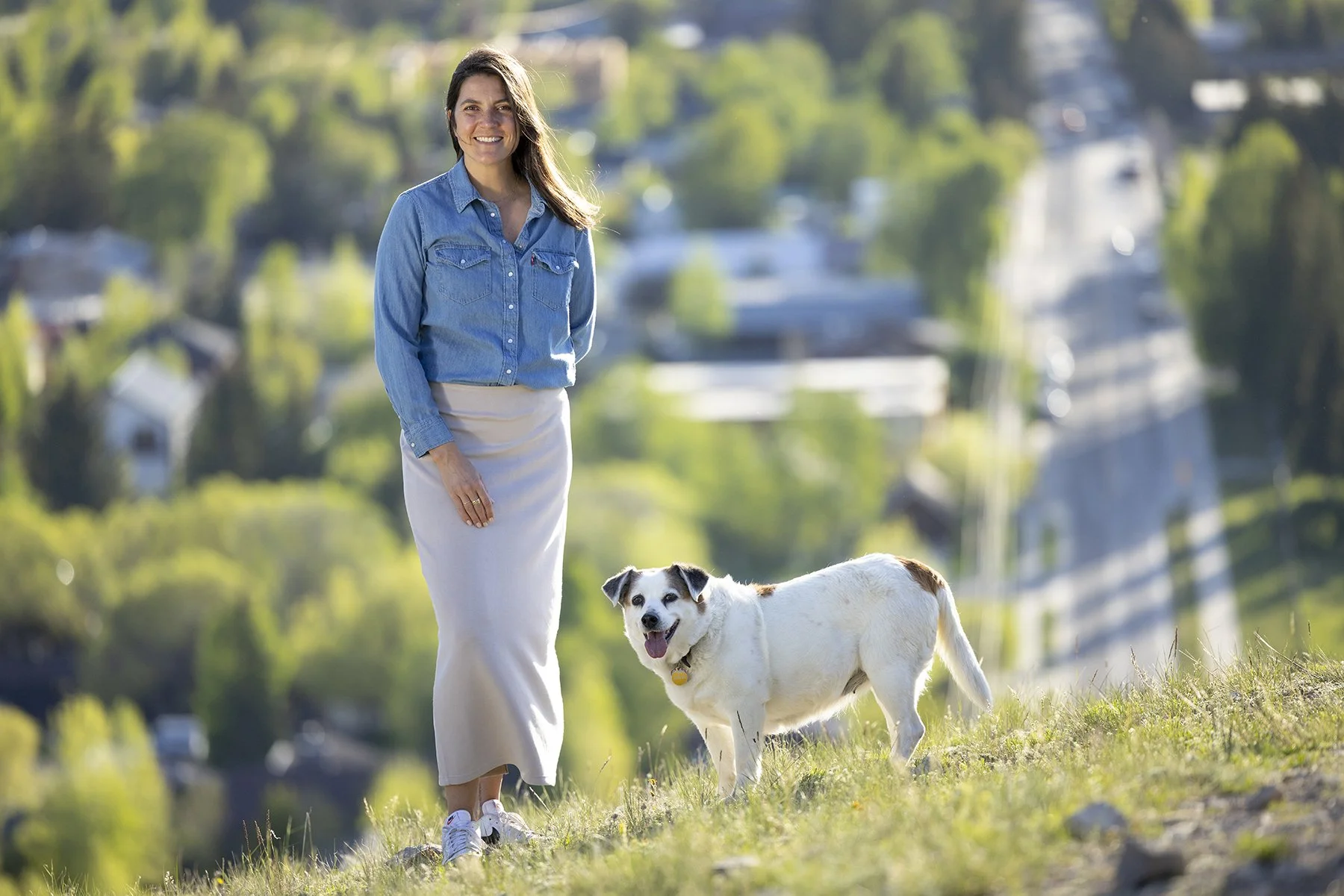 A woman walking outdoors with her dog during daytime in a grassy area with a blurred background of trees and houses.
