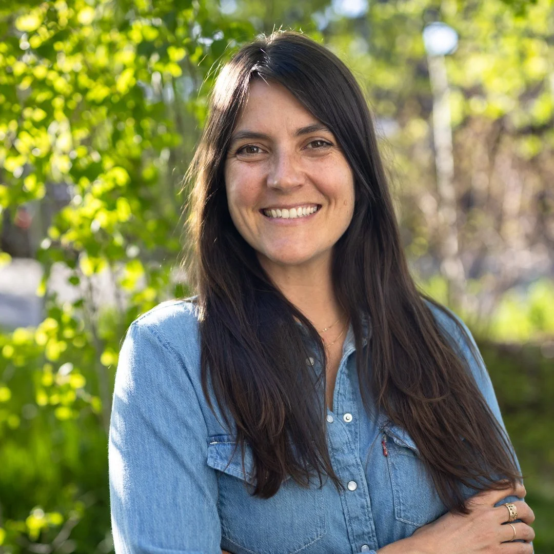 A woman with long dark hair, smiling outdoors in sunlight, wearing a denim shirt.