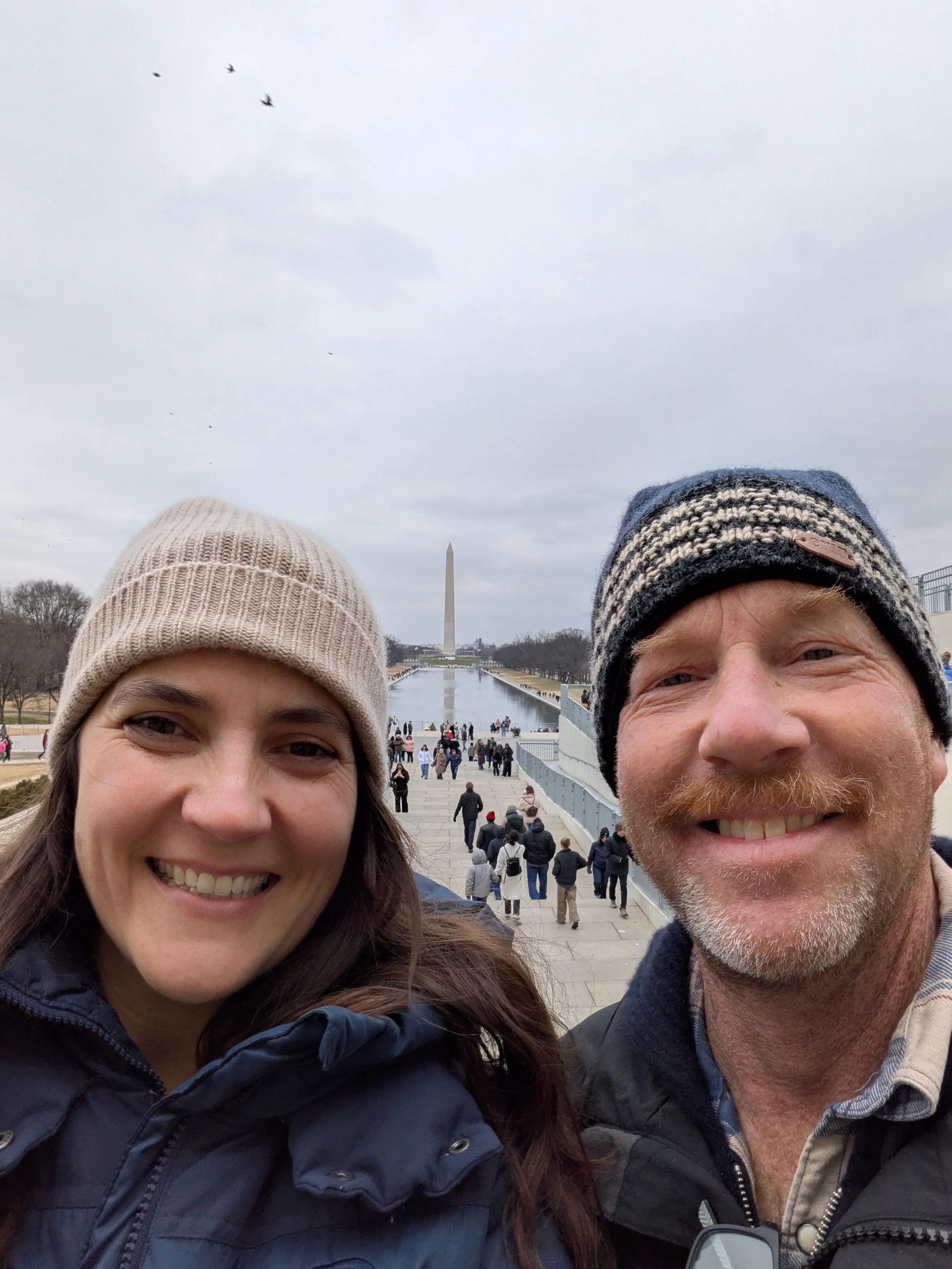 Smiling woman and man taking a selfie at the Washington Monument in Washington, D.C., with other visitors in the background.